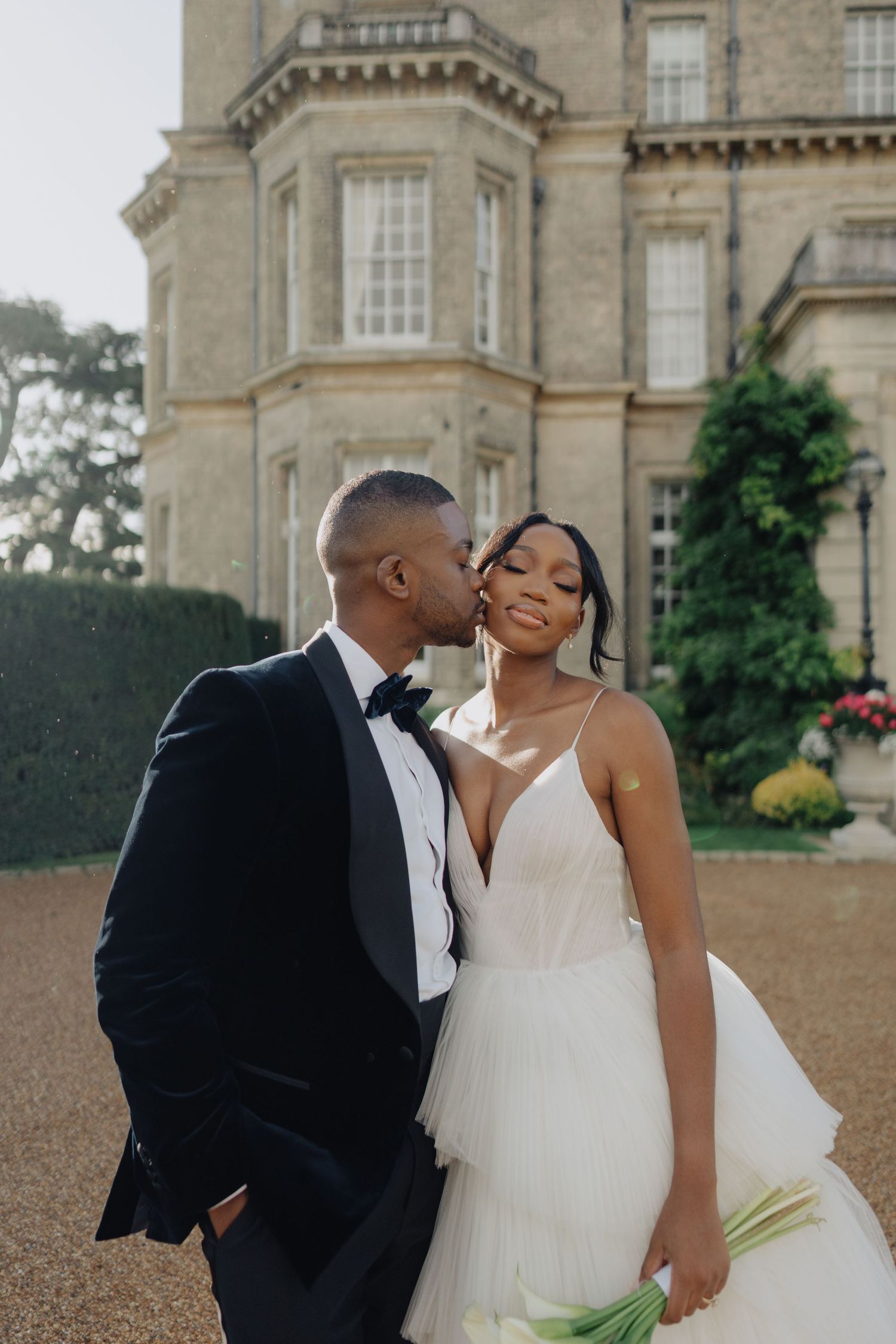 A romantic wedding couple embraces in an outdoor field setting with trees in the background.