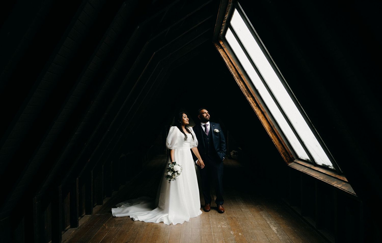 A couple in wedding attire walks together through a rustic wooden barn with sunlight streaming through windows.