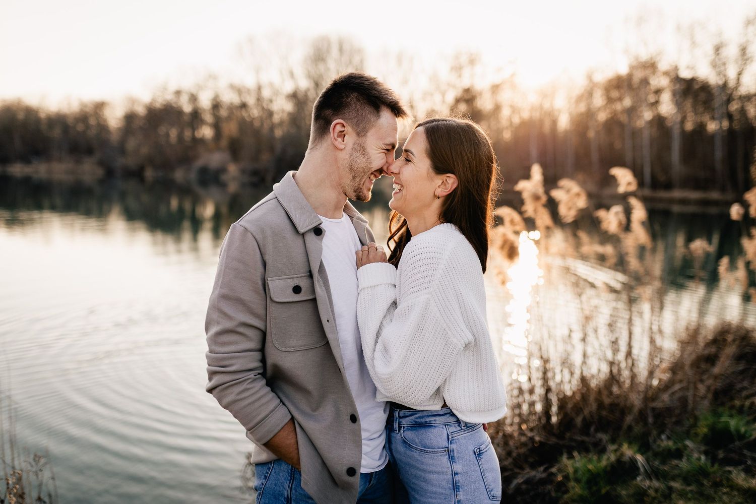 A romantic couple embraces by a serene lake at sunset, wearing casual outfits in warm golden light.
