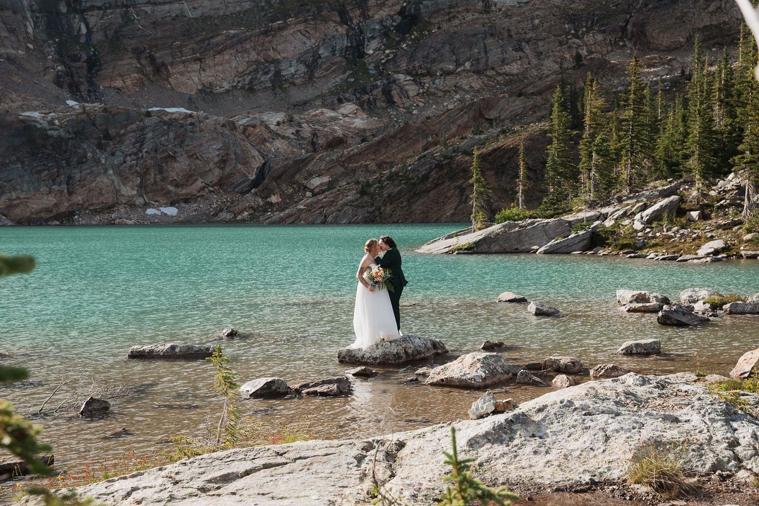 Two brides kiss on rocks beside a stunning turquoise alpine lake surrounded by mountains.