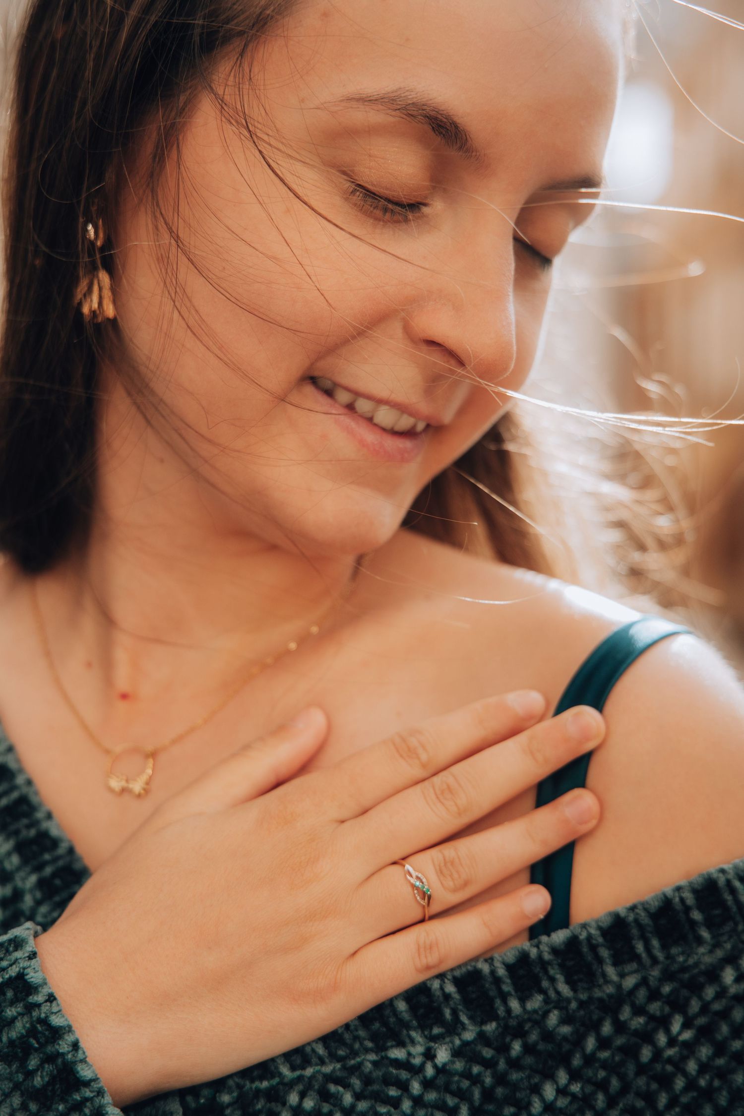 Smiling woman with eyes closed touching her shoulder wearing gold earrings and diamond ring.