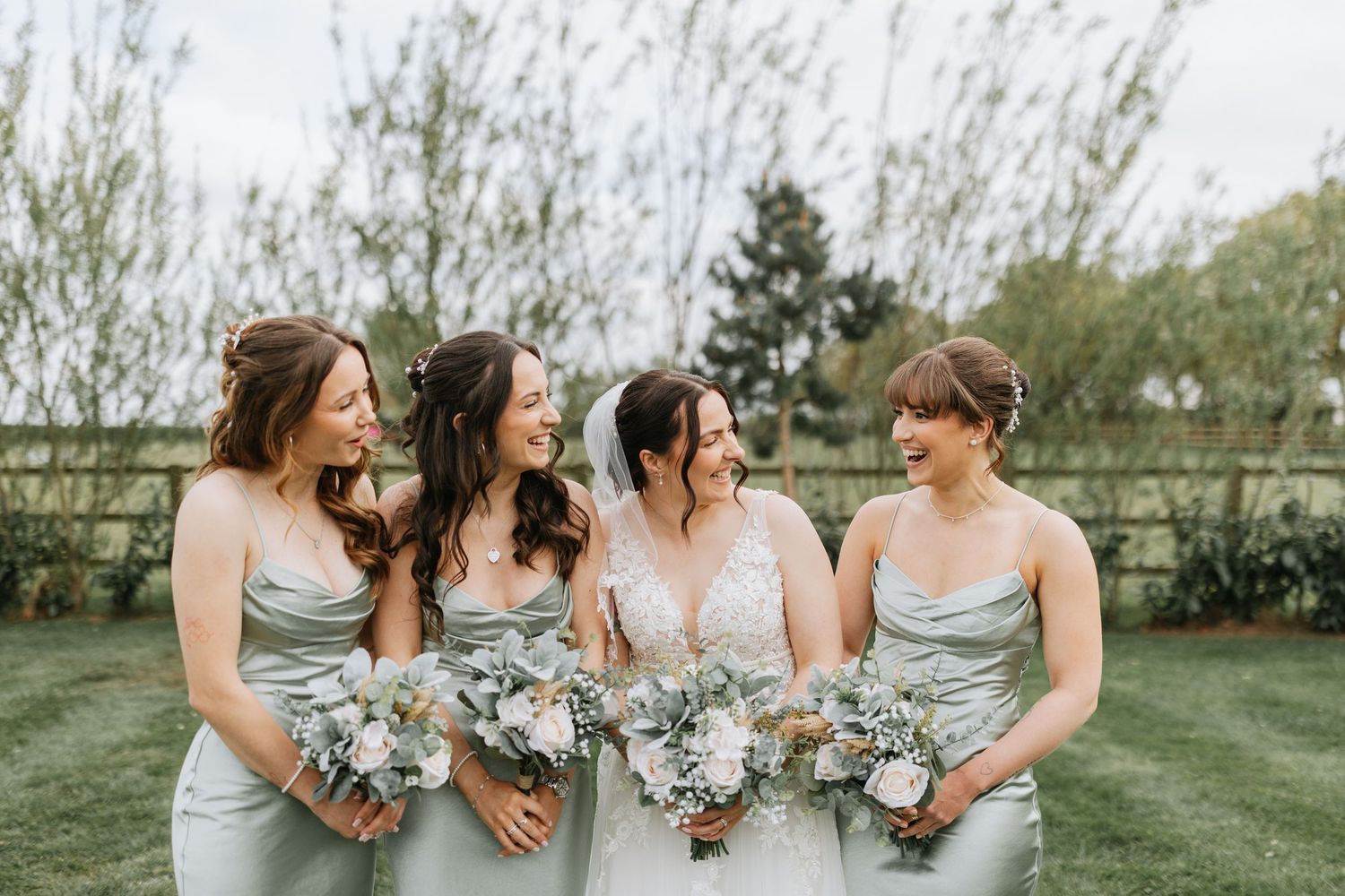 A group of bridesmaids wearing dusty blue dresses share a candid moment together in an outdoor garden setting.