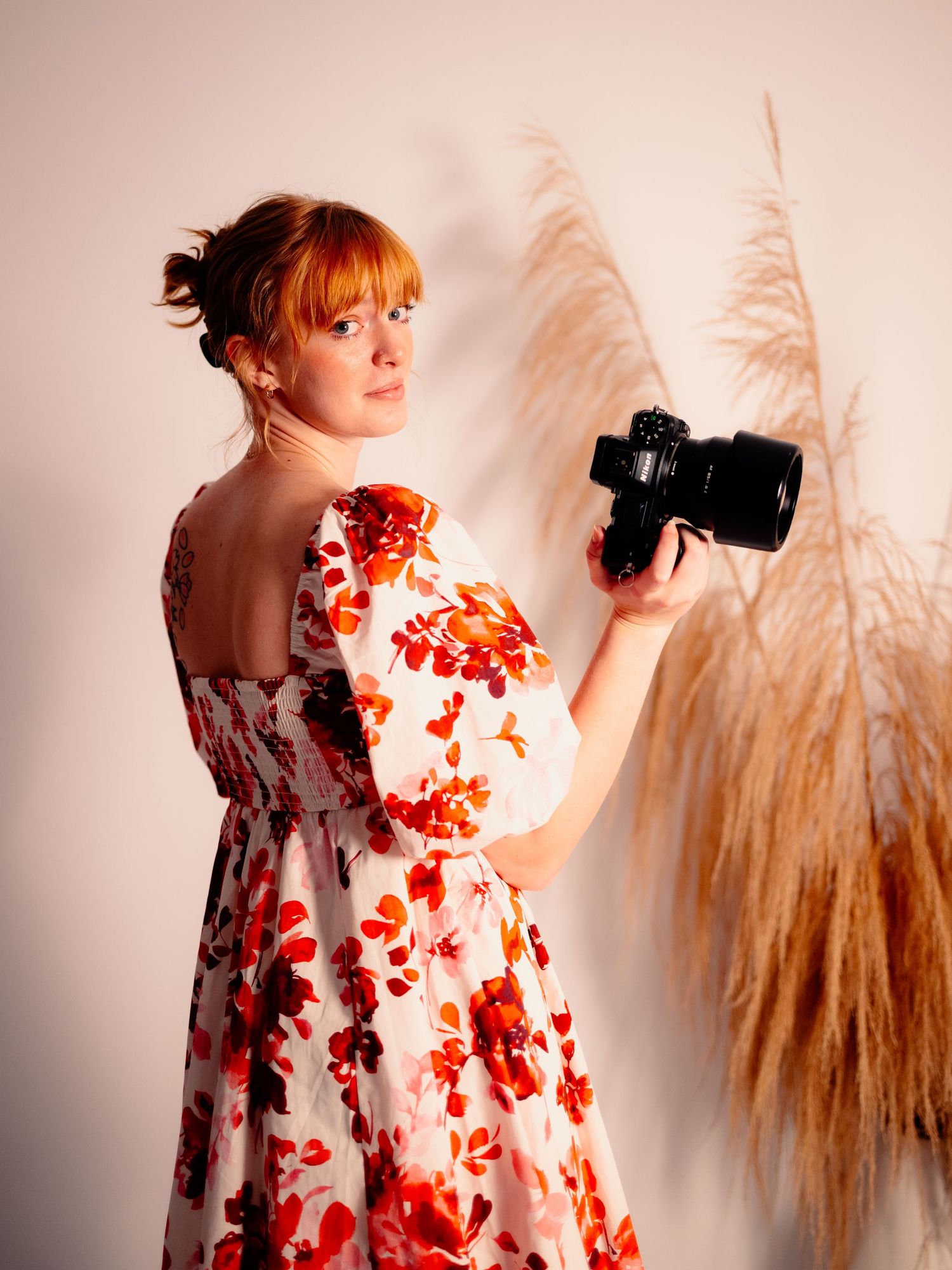 A photographer in a floral dress holds a camera against a backdrop of pampas grass and warm lighting.