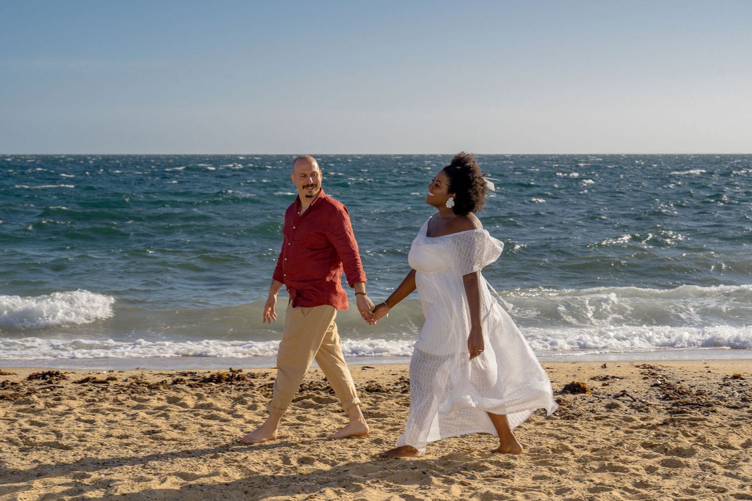 Couple walking together on a sandy beach with ocean waves in the background during sunset.