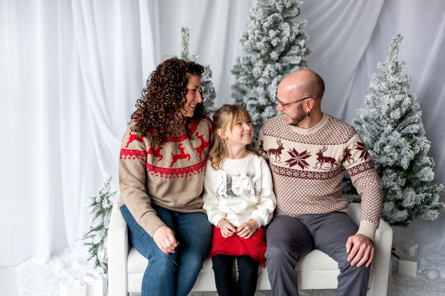 A happy family of three in matching Nordic reindeer sweaters sits together by snow-flocked Christmas trees.