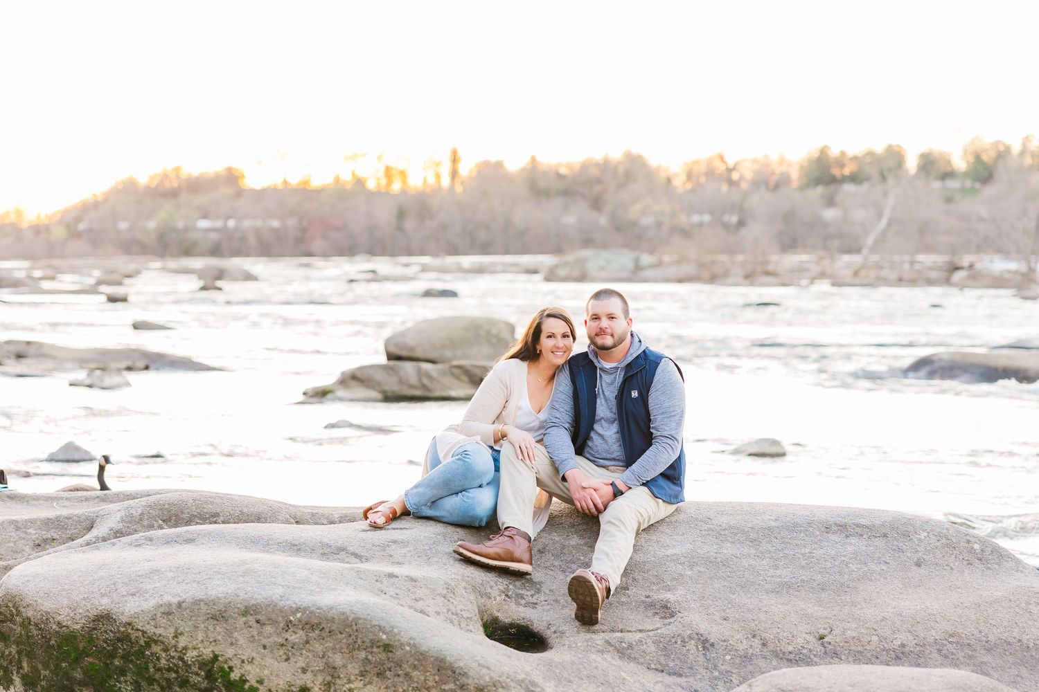 A couple sits together on large rocks at the edge of the James River at Belle Isle in Richmond during golden hour