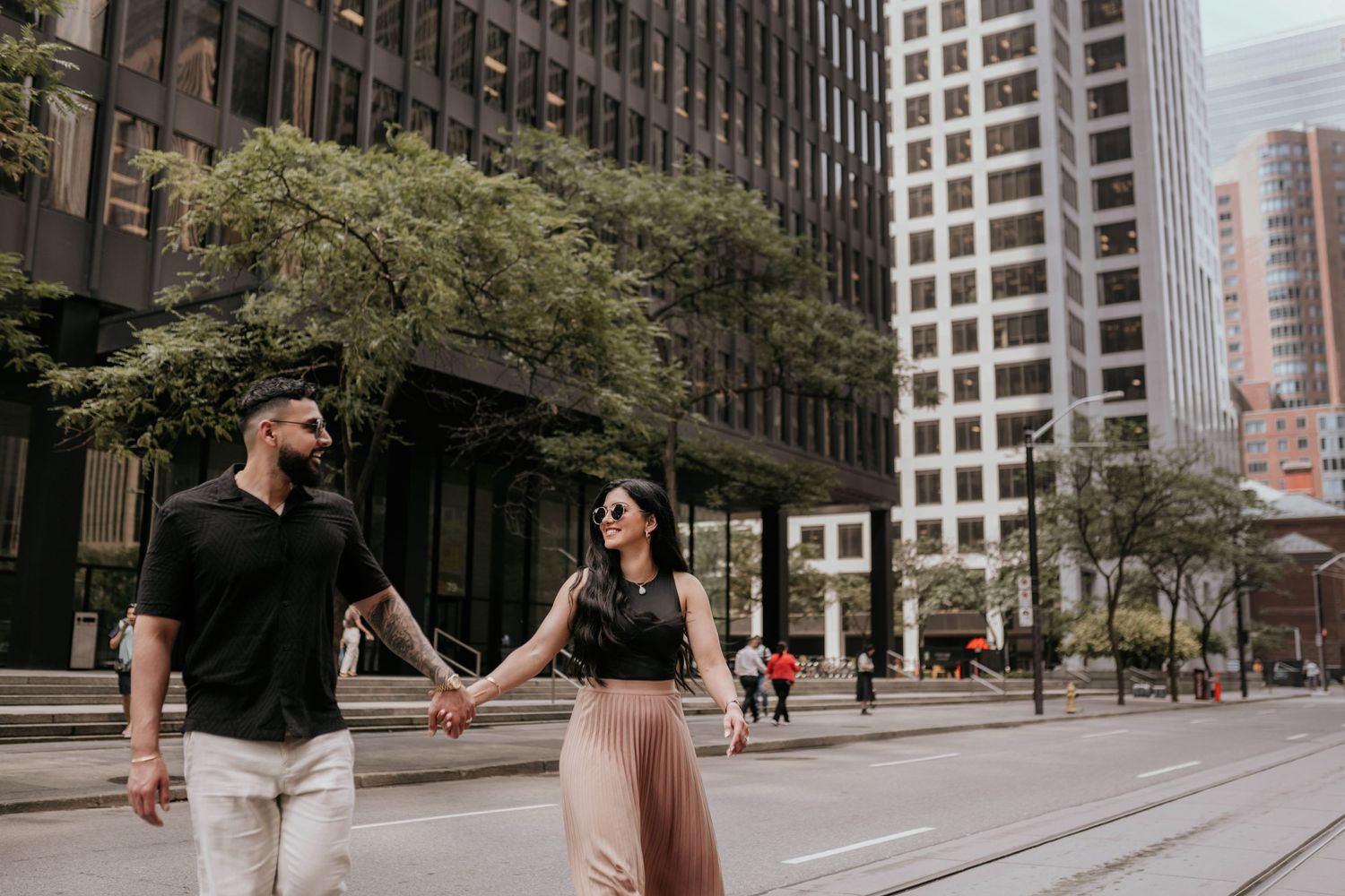A couple holds hands while walking down a city street lined with modern skyscrapers and trees in an urban setting.