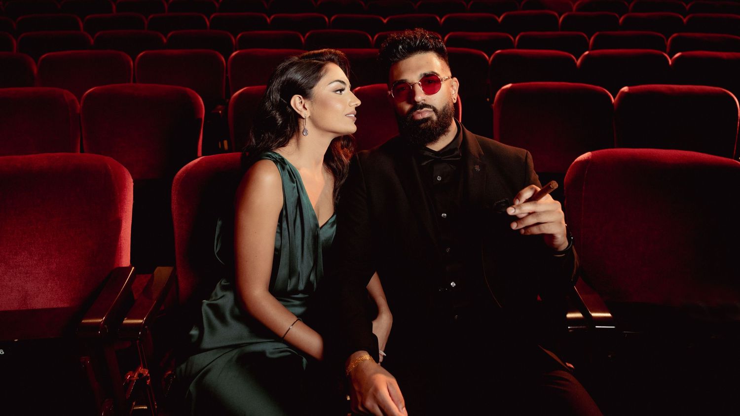 Two moviegoers sit together in red velvet theater seats having an intimate conversation in a dark cinema.