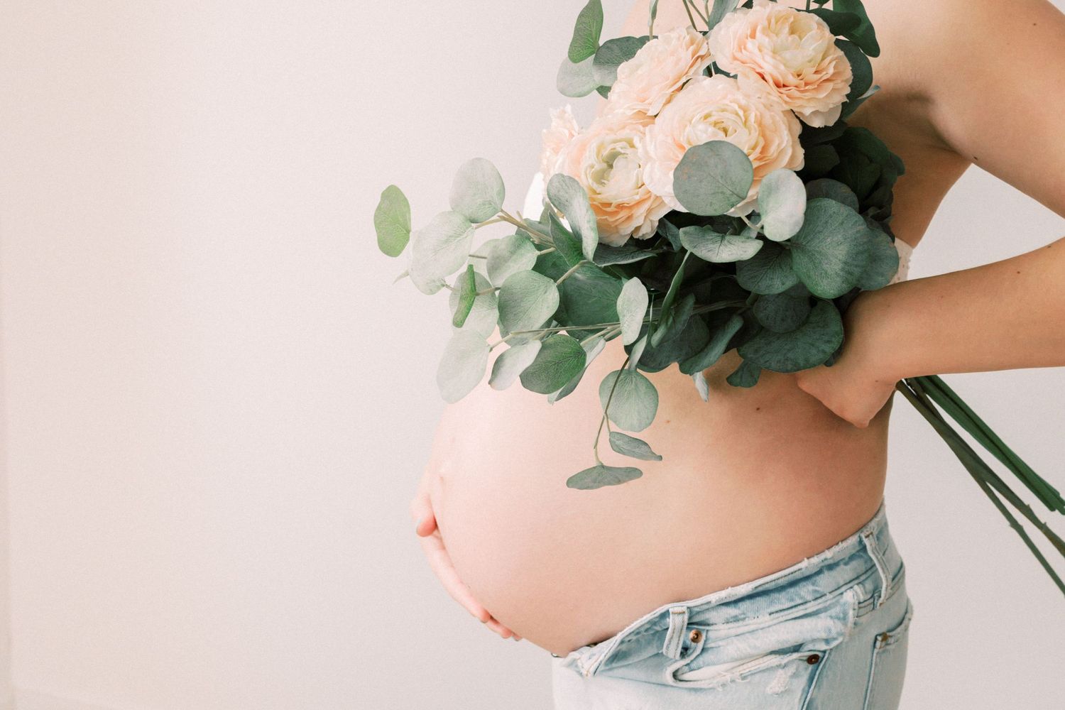 Pregnant belly shown in casual denim with a beautiful bouquet of peach roses and eucalyptus leaves against white background.
