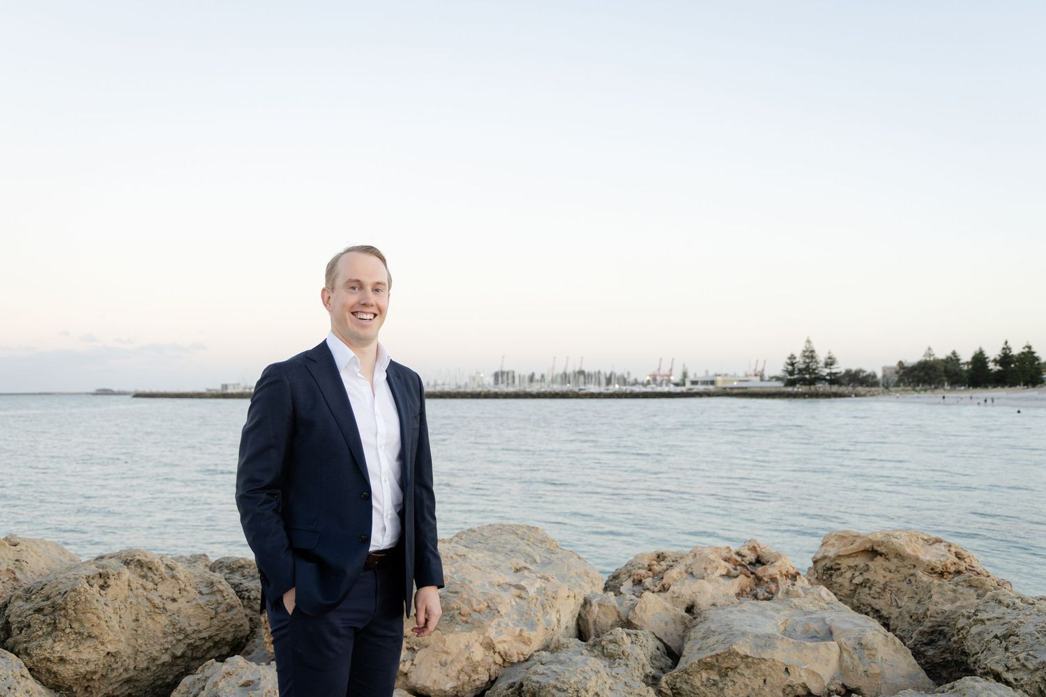 A professional in a navy suit stands on rocky waterfront with a coastal cityscape visible in the background at dusk.