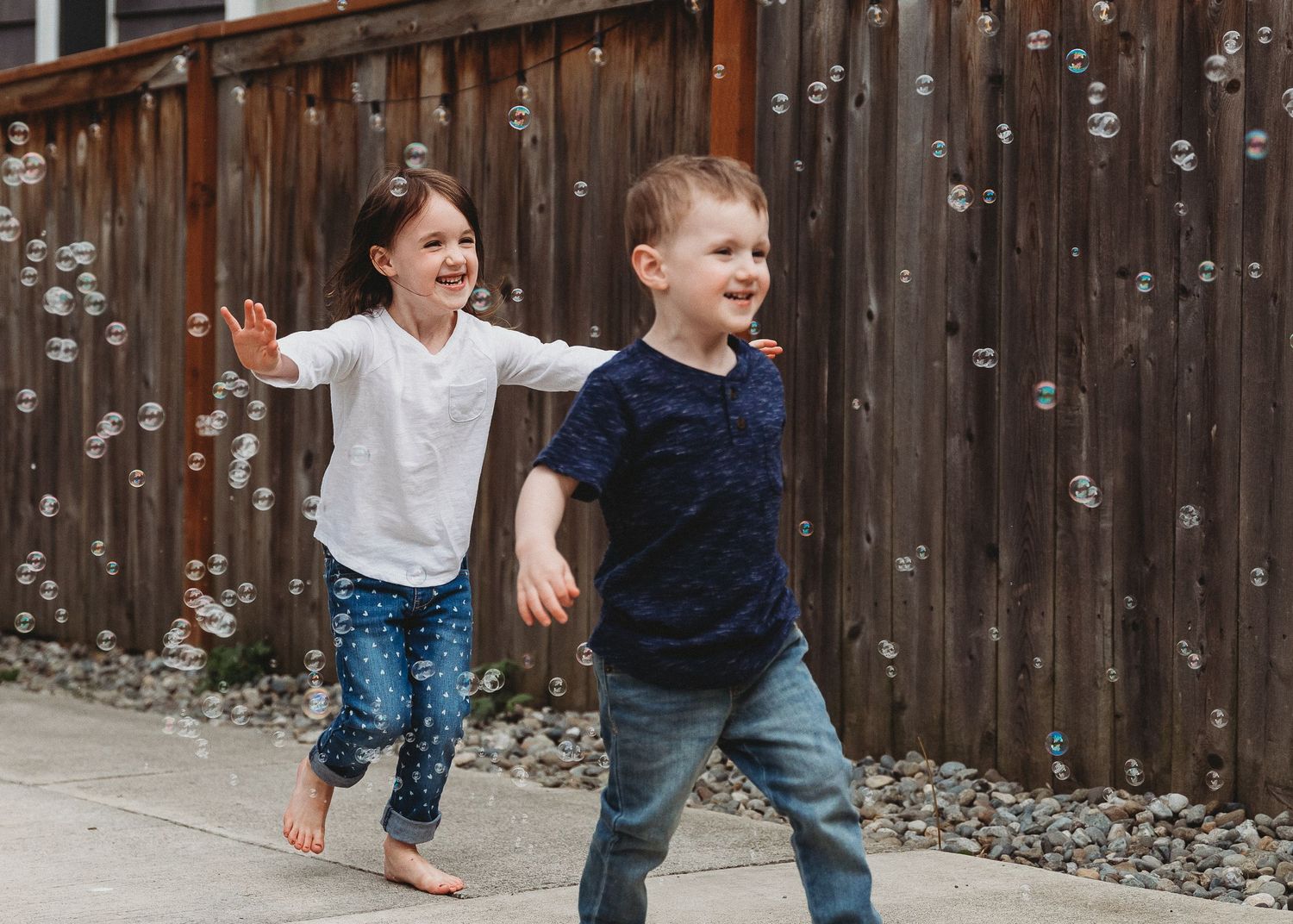 Two young children running and playing together in front of wooden fence.