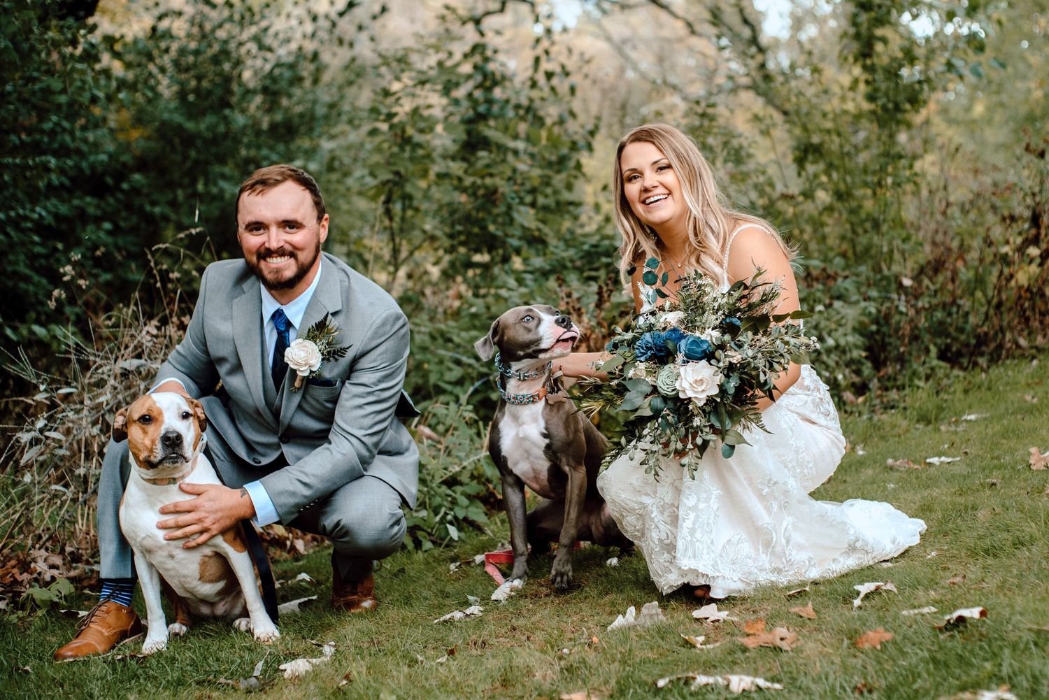 A newlywed couple poses with their dogs in formal attire during an outdoor wedding.