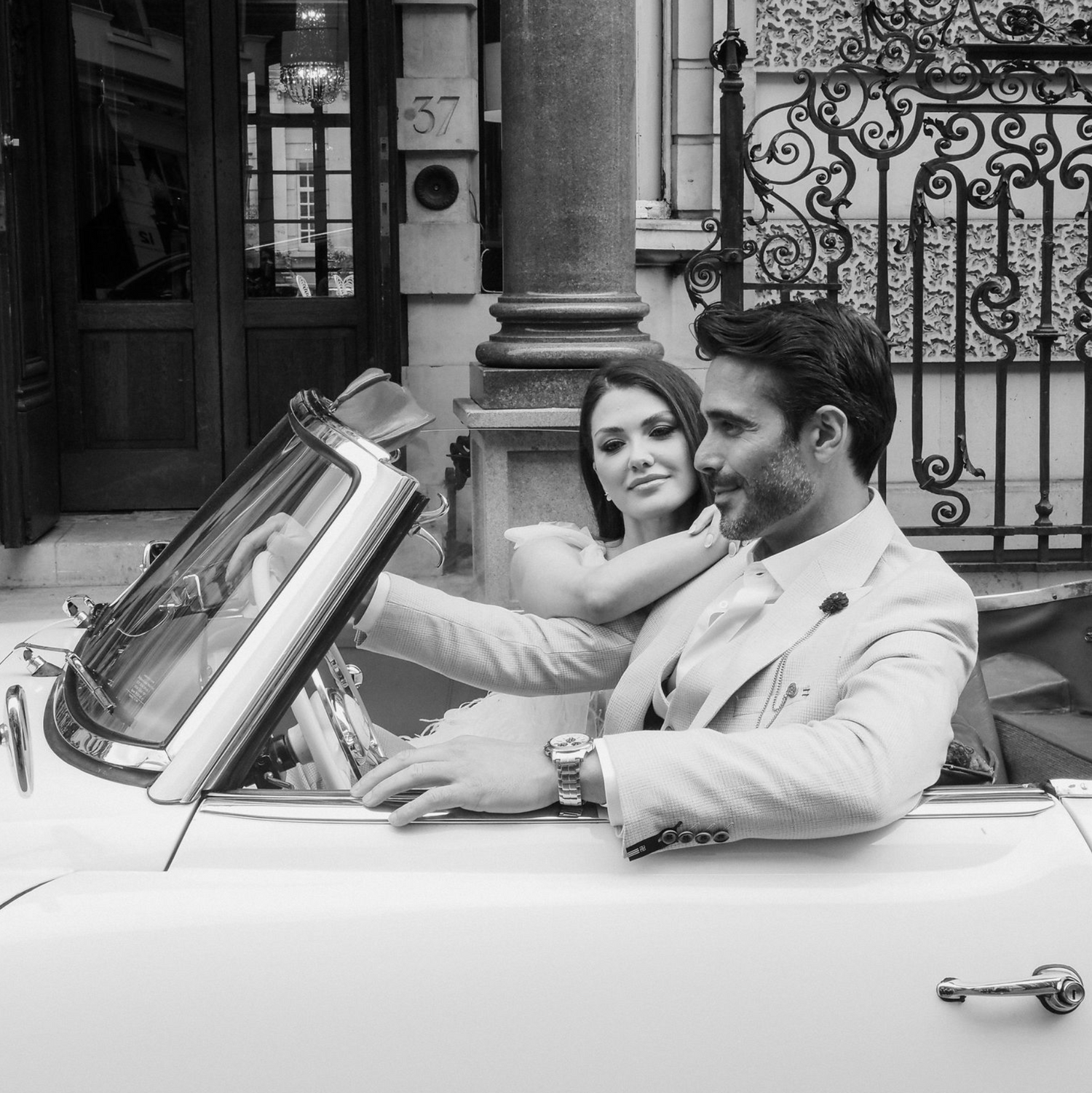 Elegant couple in vintage convertible car outside classic architecture.
