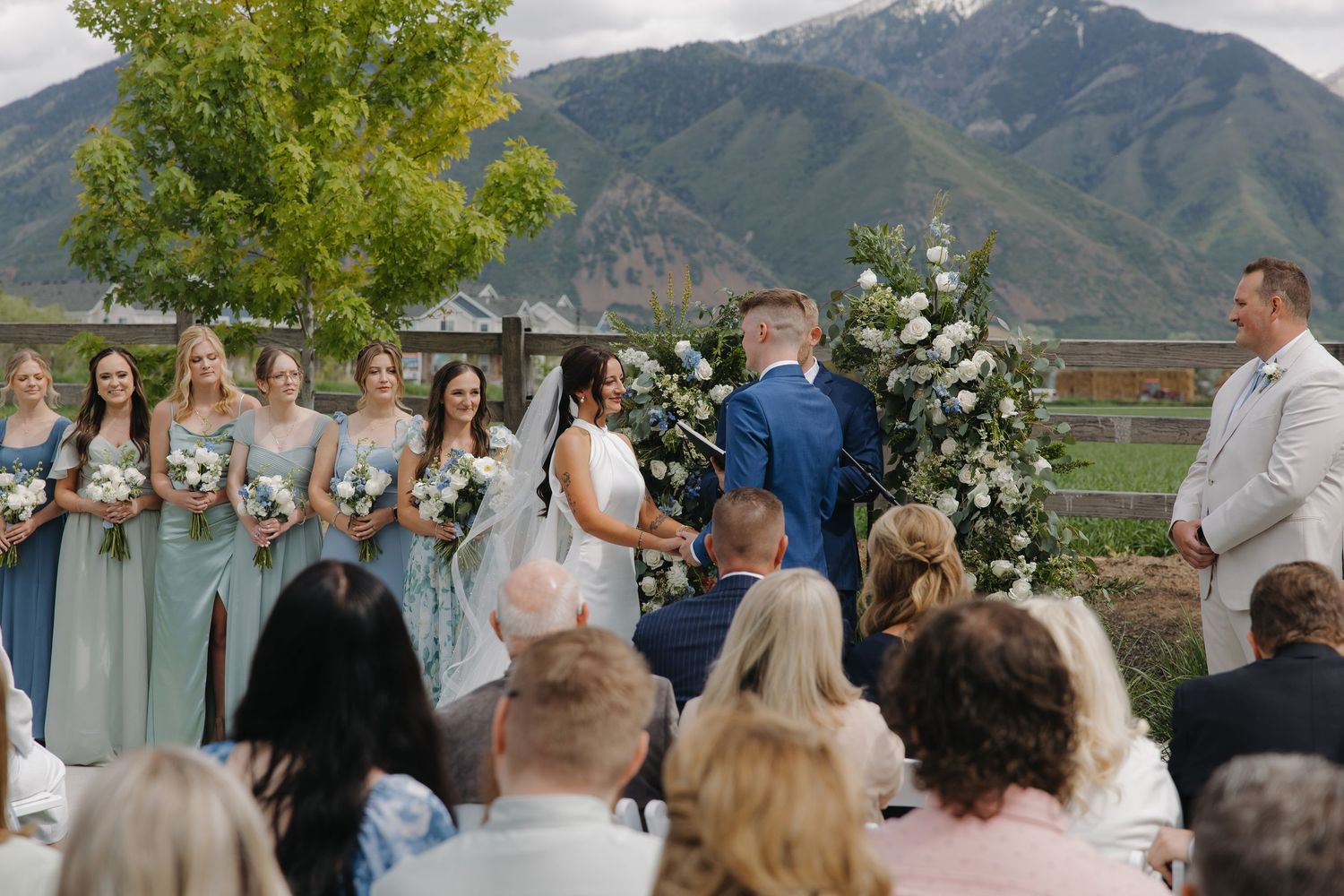 Outdoor wedding ceremony with bridesmaids in sage green dresses, floral arch, and mountain backdrop.