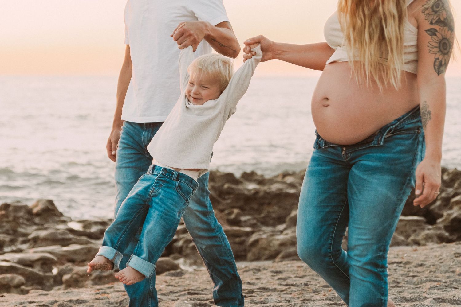 Family playing together on a beach at sunset with a young child jumping while holding hands on the sandy shore.