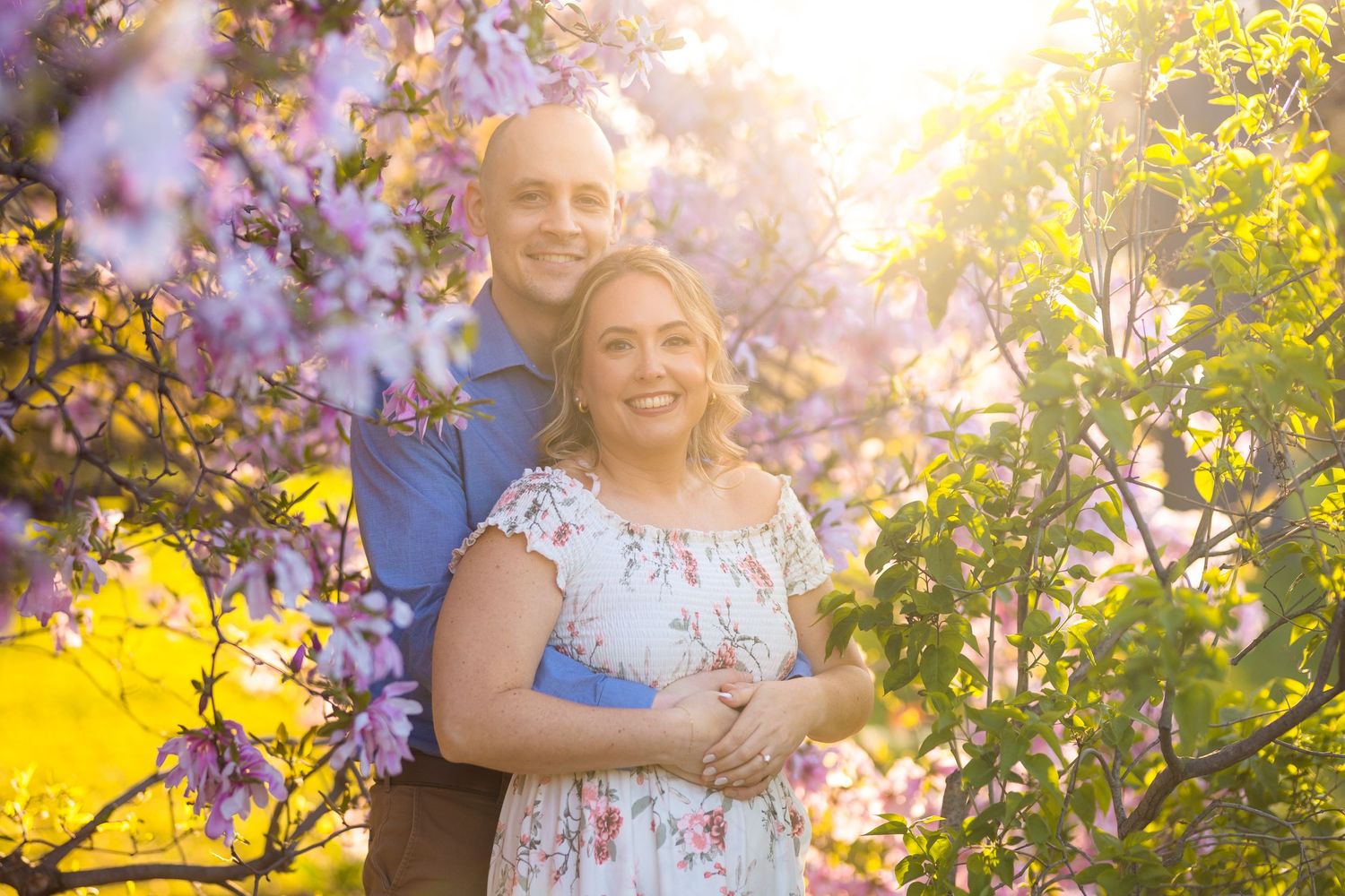 A couple stands among blooming spring flowers with golden sunlight filtering through trees in a romantic outdoor setting. Ottawa Wedding