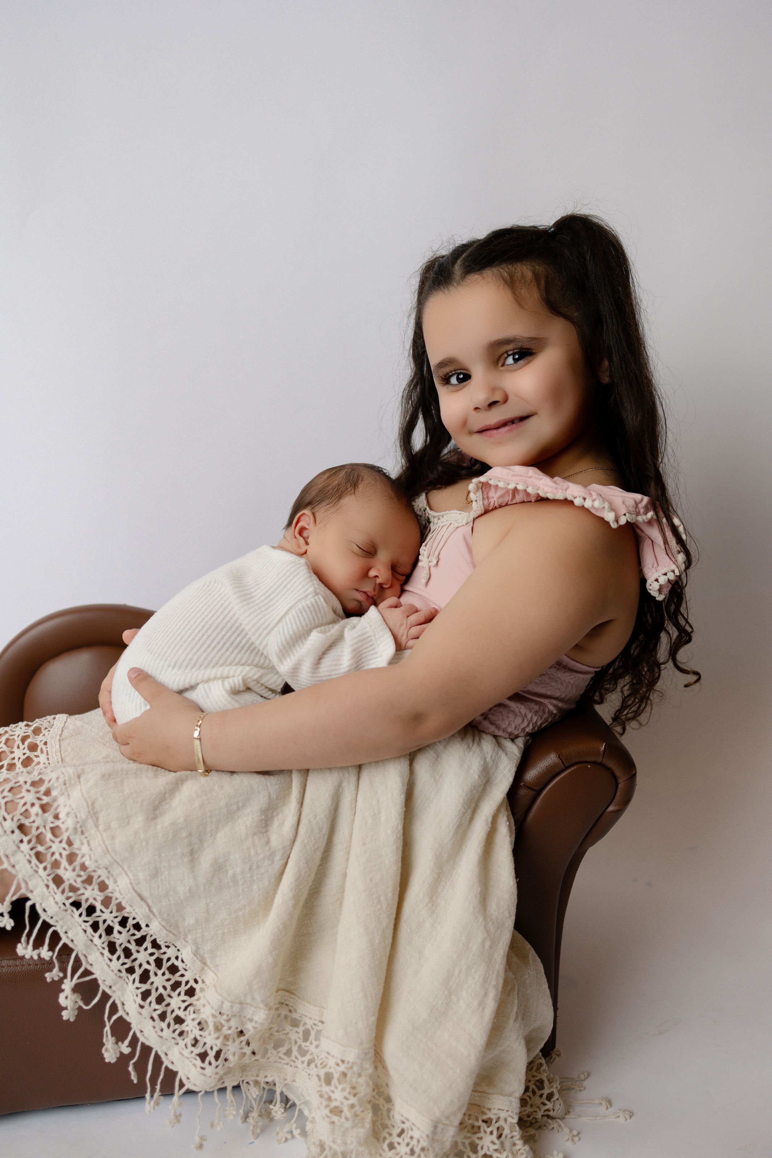 A young girl in a pink dress sits in a brown chair while holding a sleeping newborn wrapped in cream-colored lace blanket.