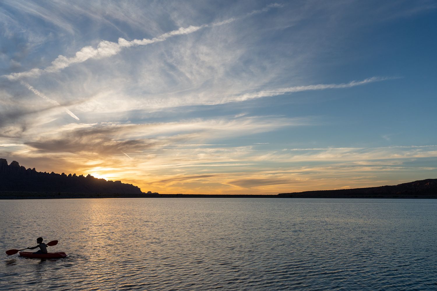 Silhouette of kayaker on calm lake waters during golden sunset.