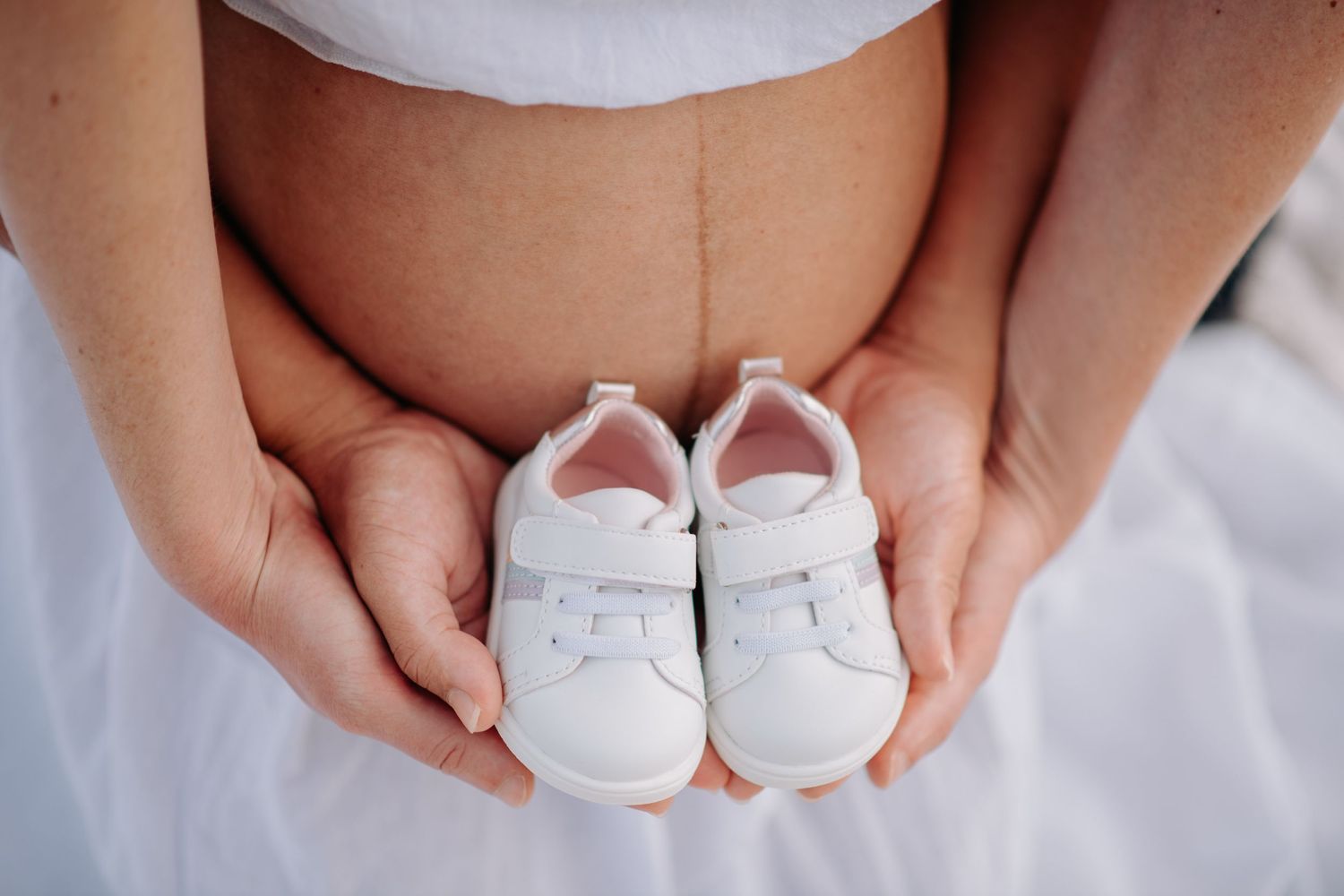 Hands holding tiny white baby shoes against pregnant belly in tender maternity moment.
