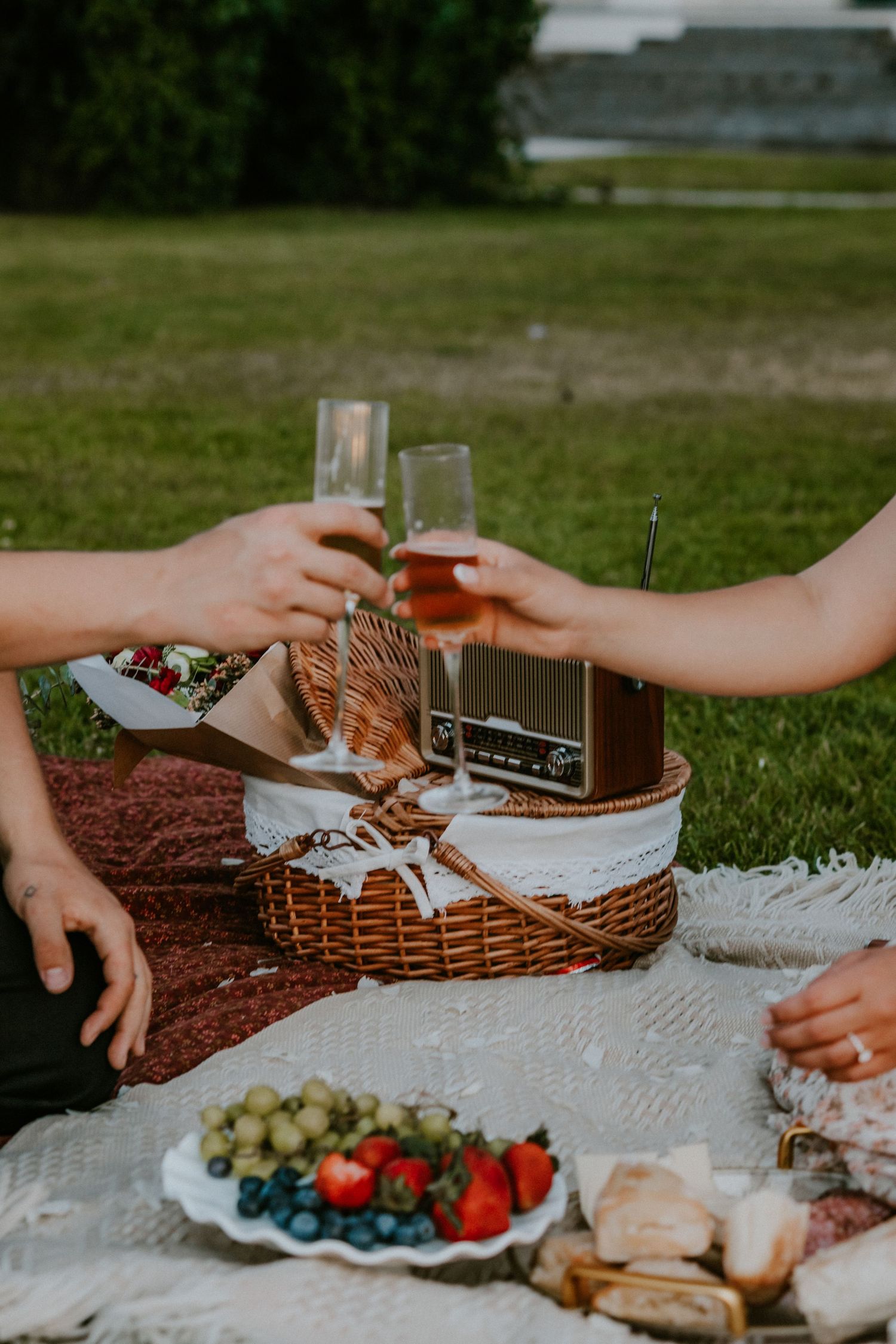 Two people toasting champagne glasses over a picnic basket with fresh fruit and bread on a blanket outdoors.