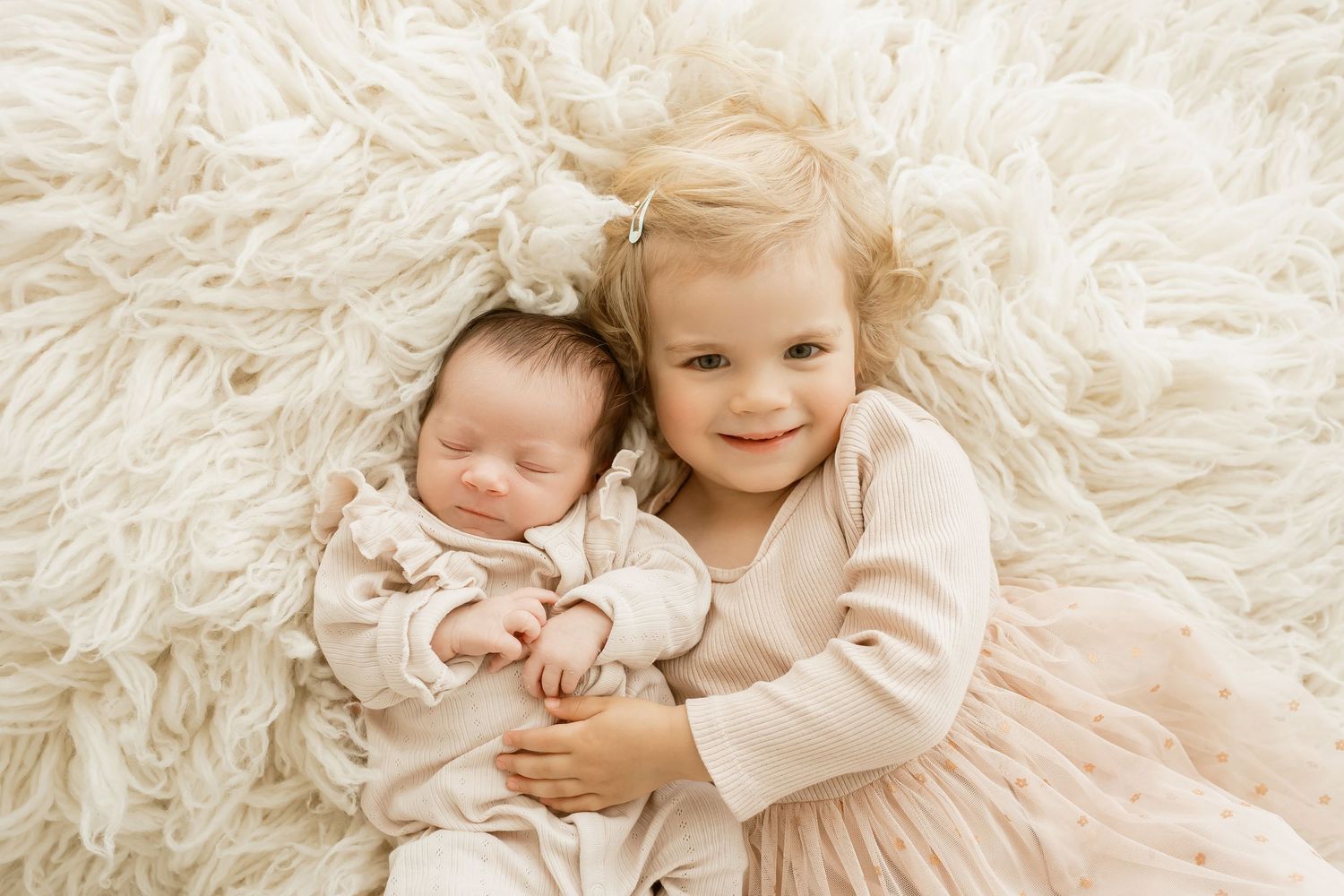 Two children in soft pink attire lying on white fluffy rug with the older sibling embracing the younger one.