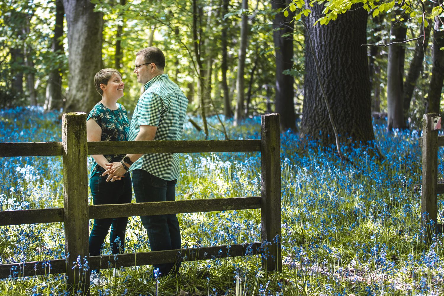 A couple stands by a wooden fence in a spring forest filled with blooming blue wildflowers on a sunny day.