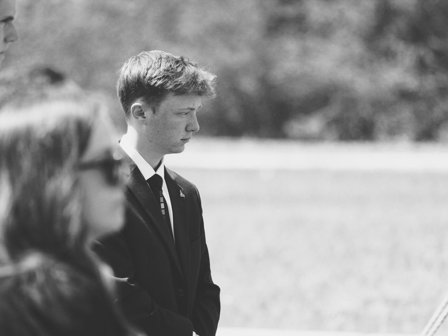 A black and white photo of a young man quietly watching the graveside services.