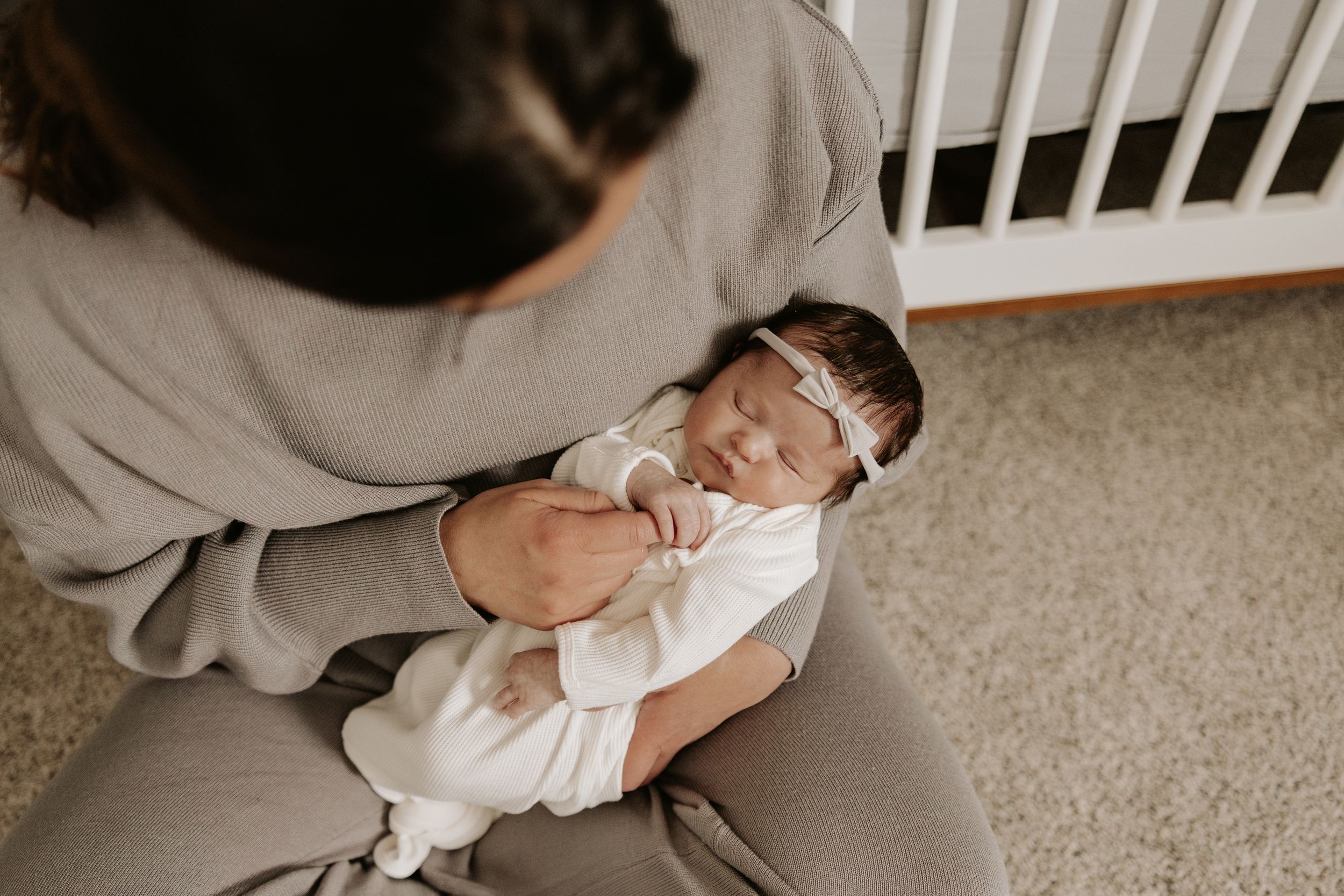 A peacefully sleeping newborn baby in white clothing rests in someone's arms on a carpeted floor.