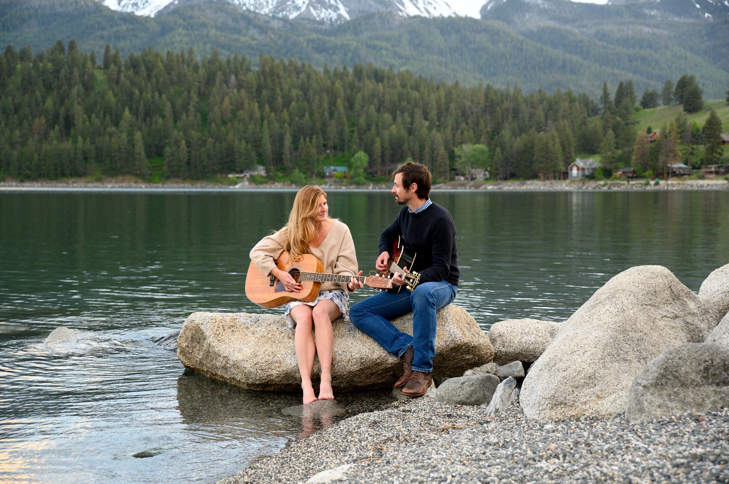 Couple sits together on rocks by a mountain lake playing guitar.