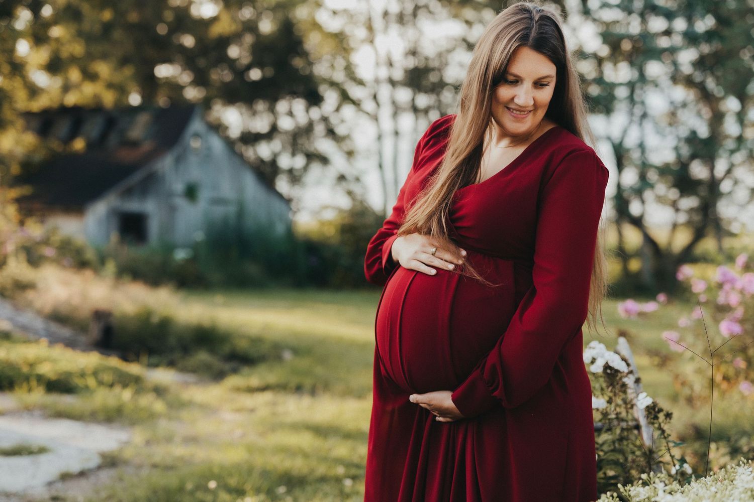 Expectant mother in a burgundy dress smiles while cradling her belly in a sunlit garden setting.