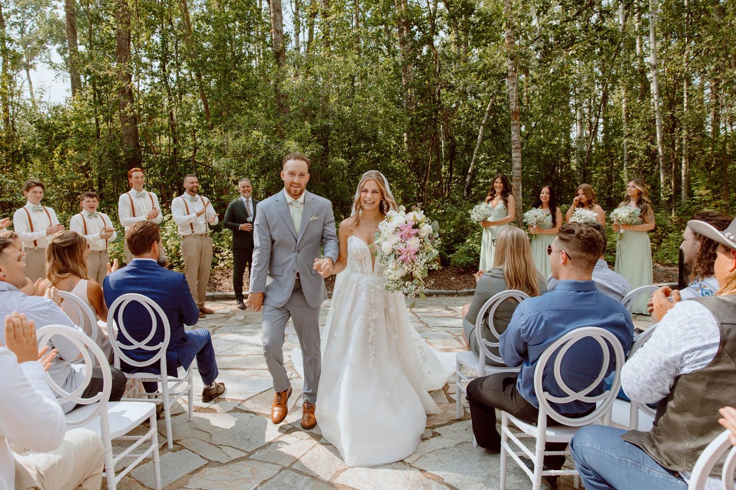 Outdoor forest wedding ceremony with white chairs and wedding party in light formal attire.