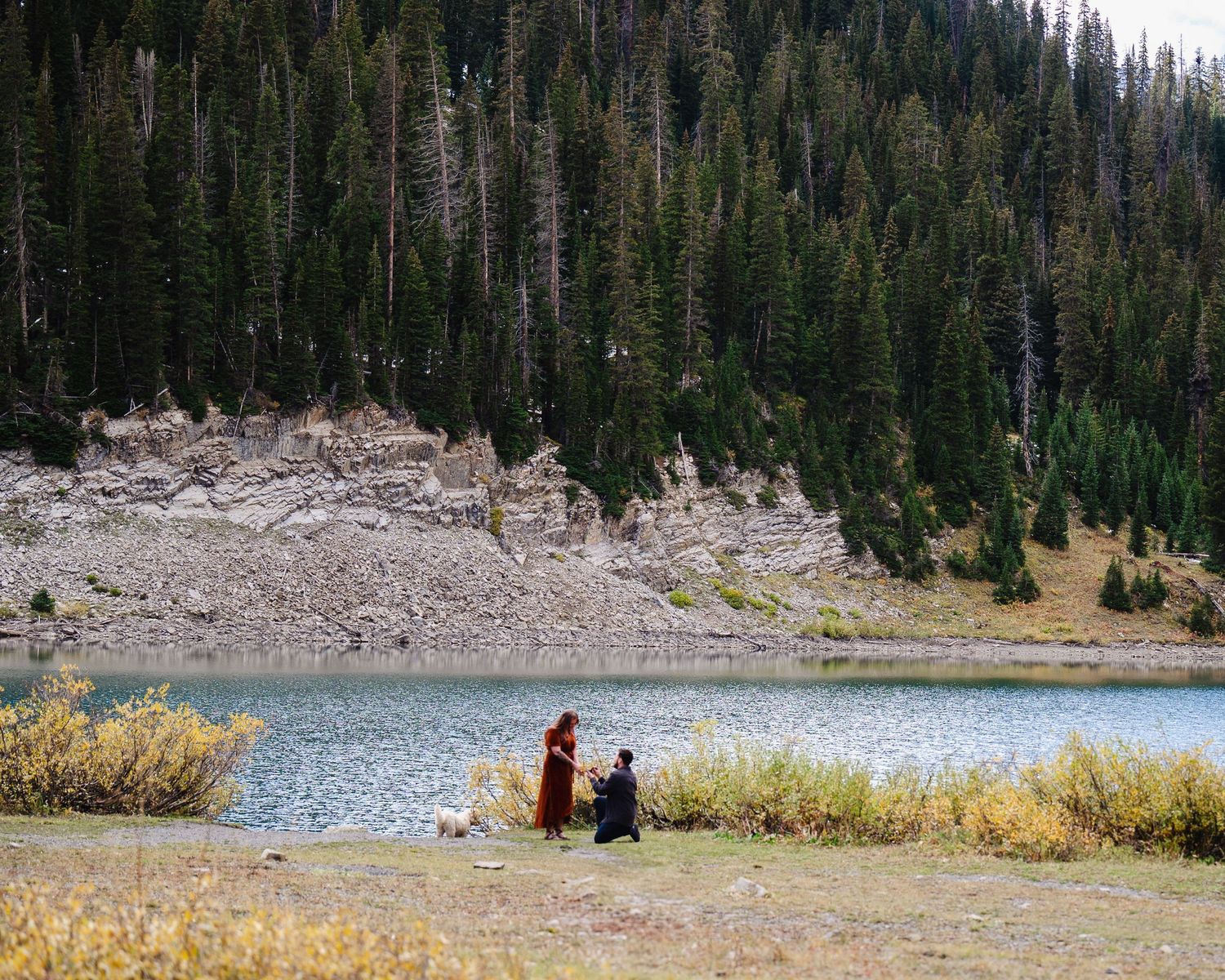 Two people sit by a serene mountain lake surrounded by evergreen forest and rocky terrain.