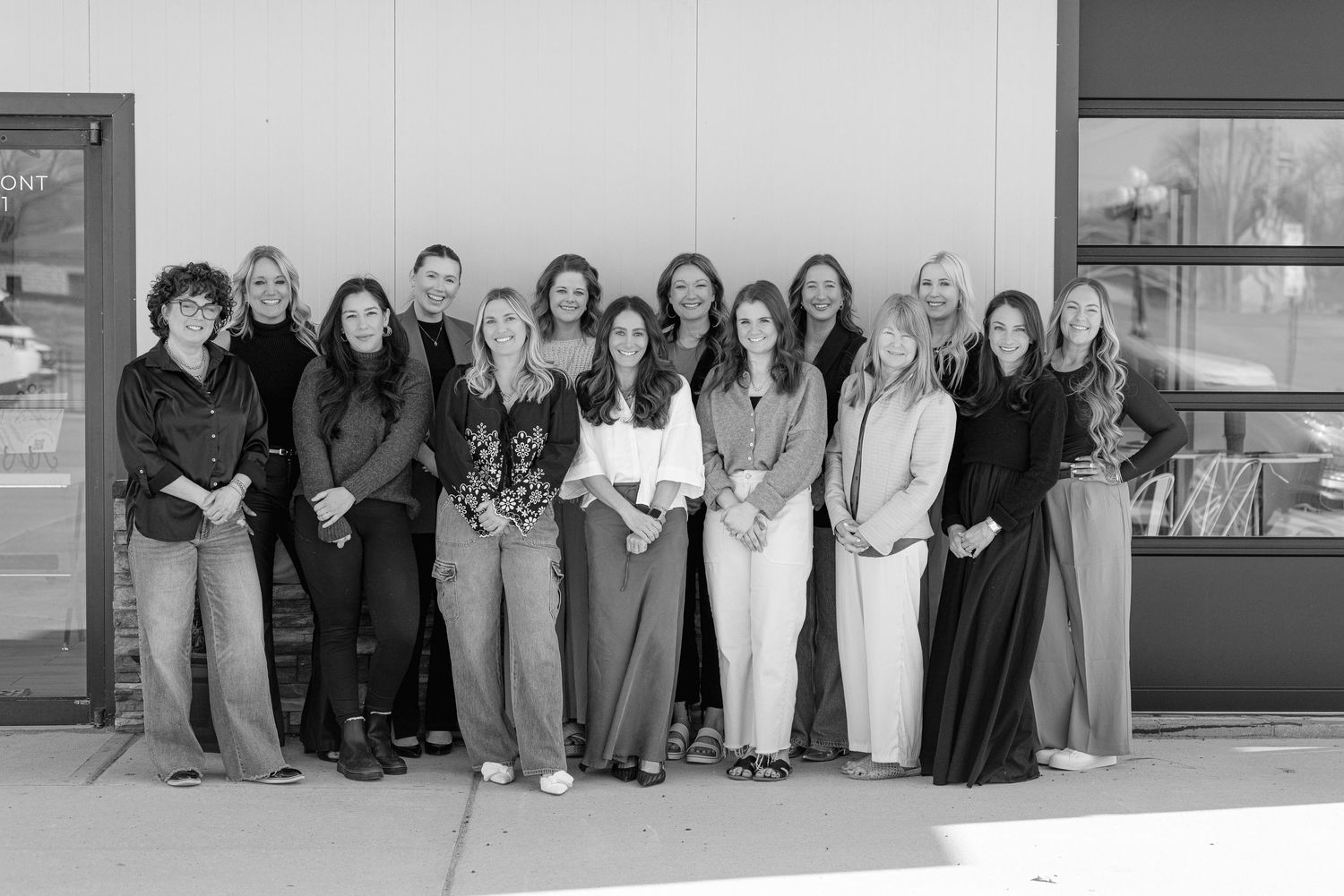 A professional group of women wearing business attire stands together in a line outside a building in black and white.
