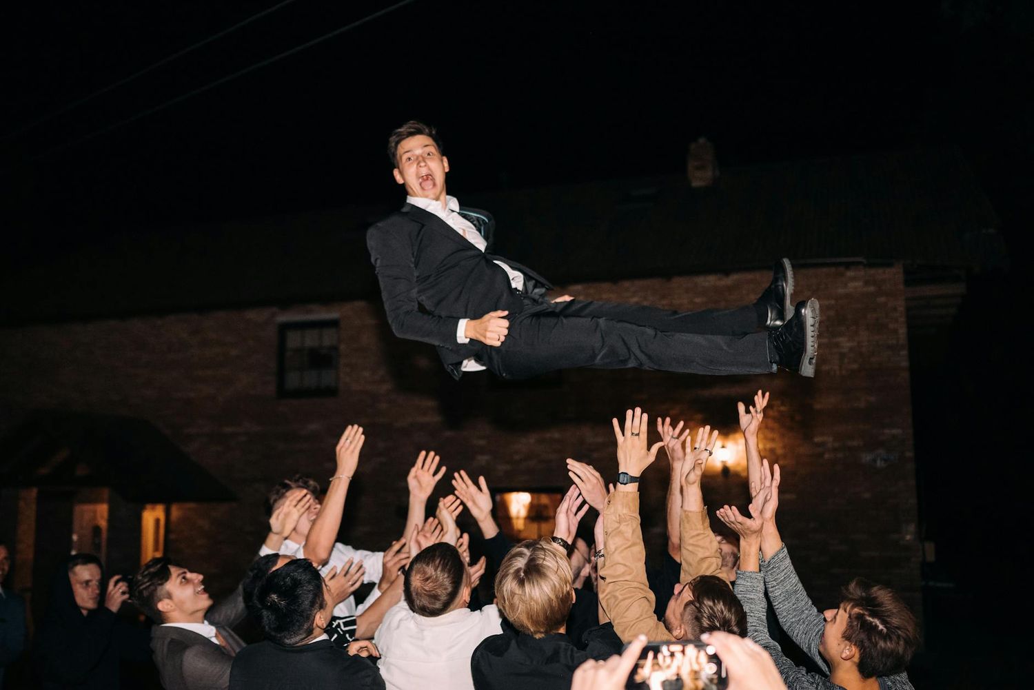 A crowd lifts and supports a person in formal attire at a wedding party.