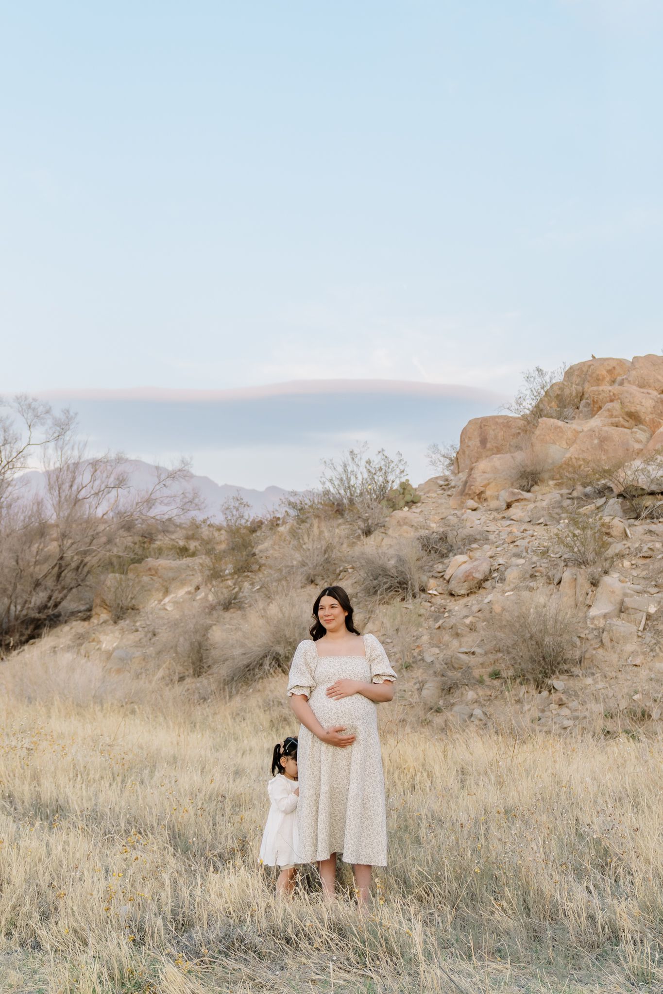 A pregnant figure in a white dress stands with a small child in a scenic desert mountain landscape at sunset, in El Paso TX