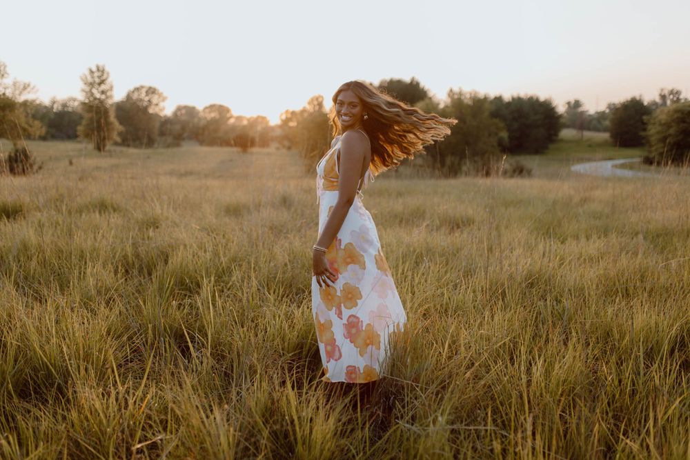 Someone in a flowing floral dress twirls in a golden sunlit meadow at sunset.