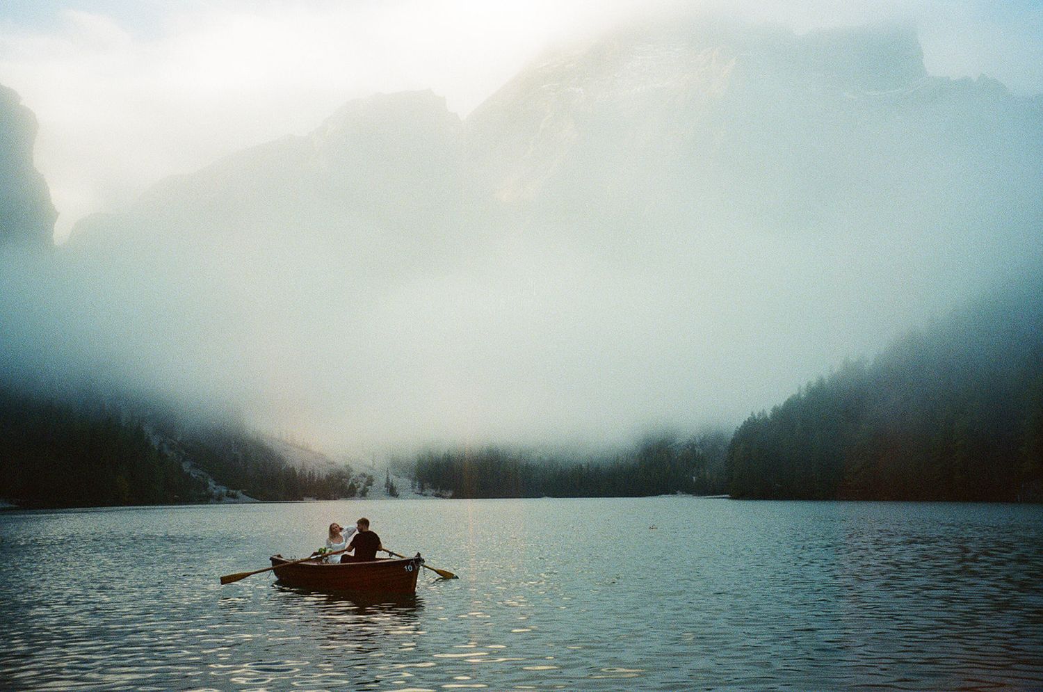 People rowing a small boat on a misty mountain lake surrounded by evergreen forests at dawn.