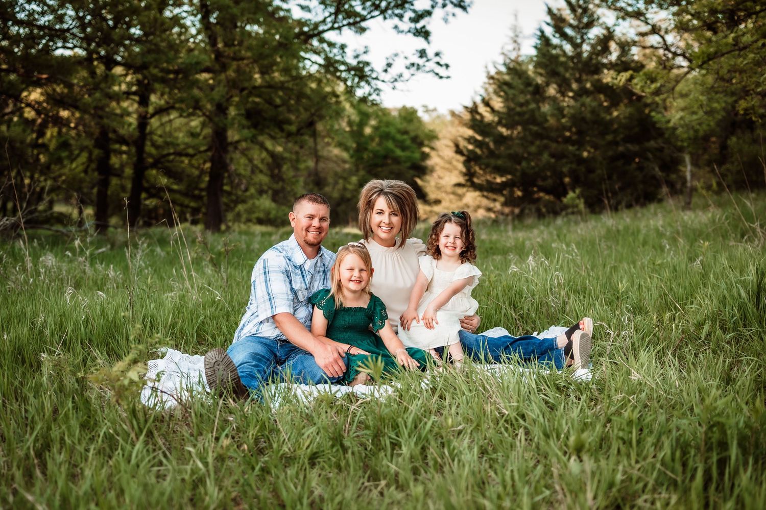 A family sits together on a blanket in tall grass with evergreen trees in the background during golden hour.