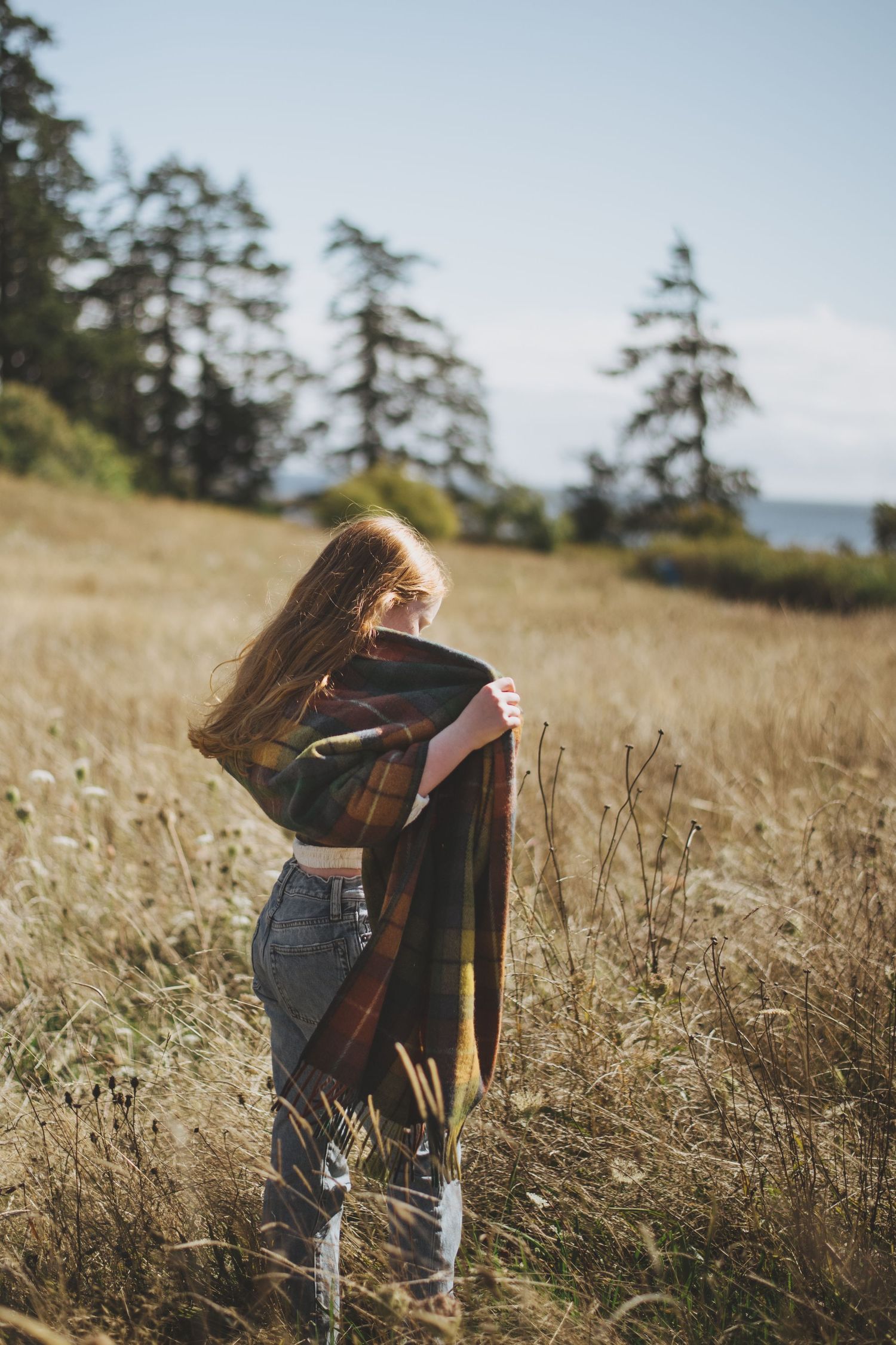 A young girl with long red hair stands in a golden grassy field, wrapped in a traditional tartan wool blanket.