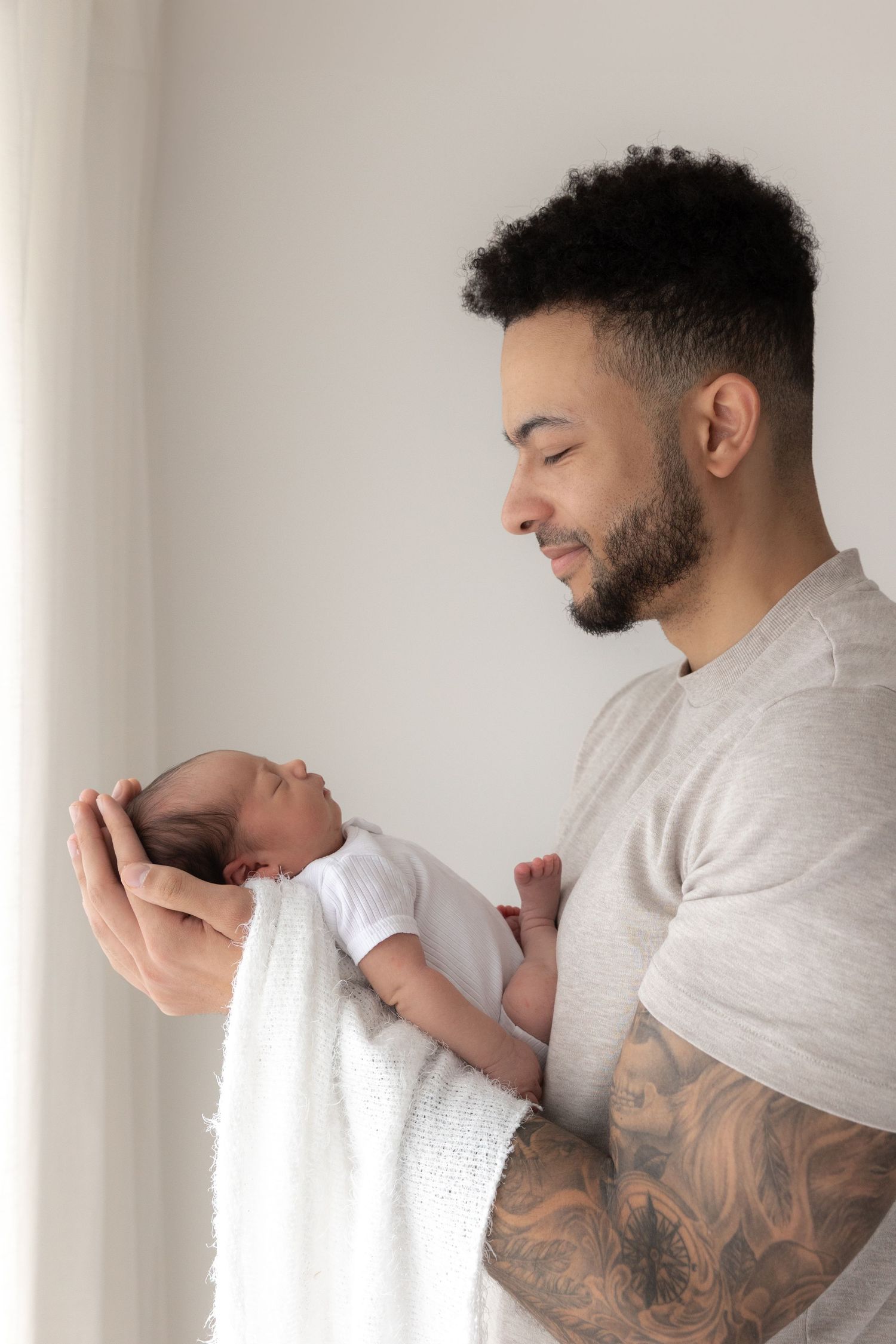 A tender moment between a father cradling his newborn baby while standing against a white wall, showing detailed arm tattoos.