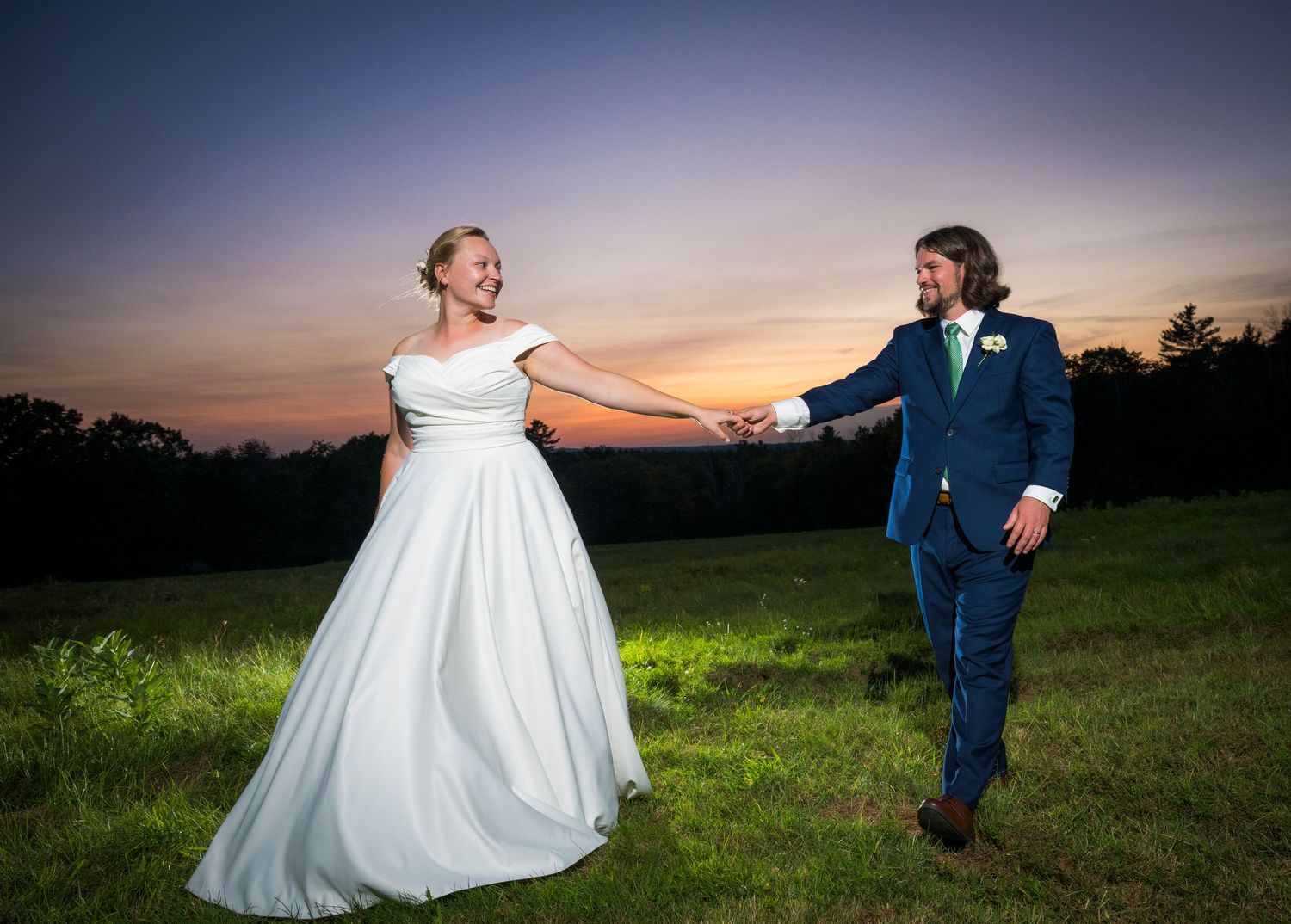 A couple in wedding attire dances playfully at sunset in an open field.