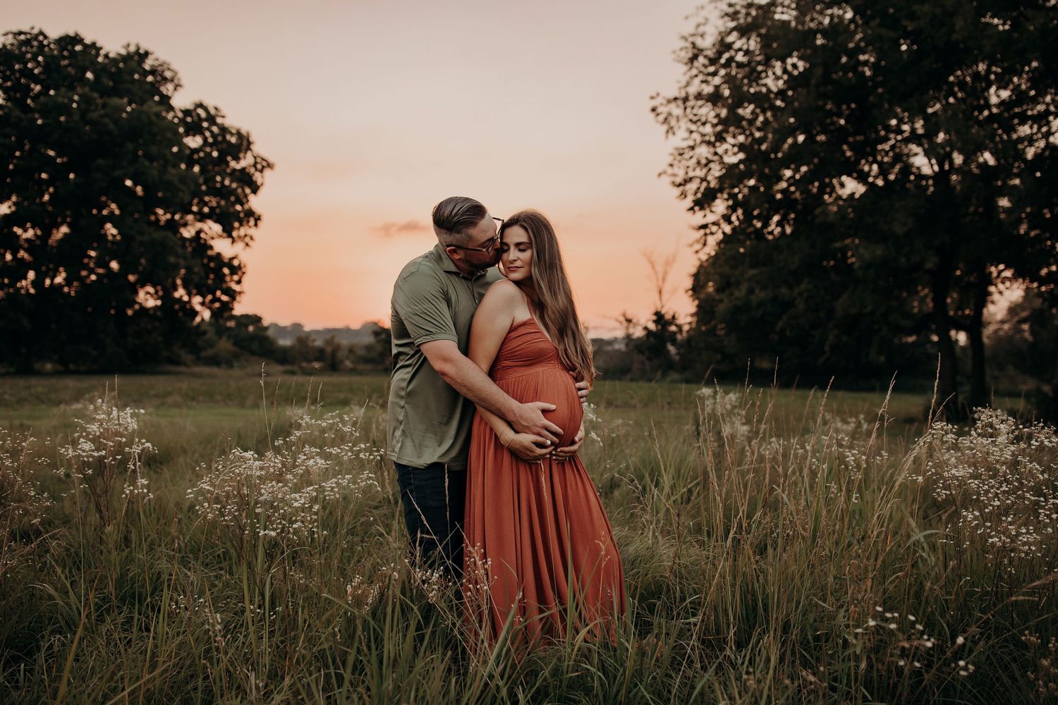 A couple embraces in a meadow at sunset with wildflowers and trees silhouetted in the background.