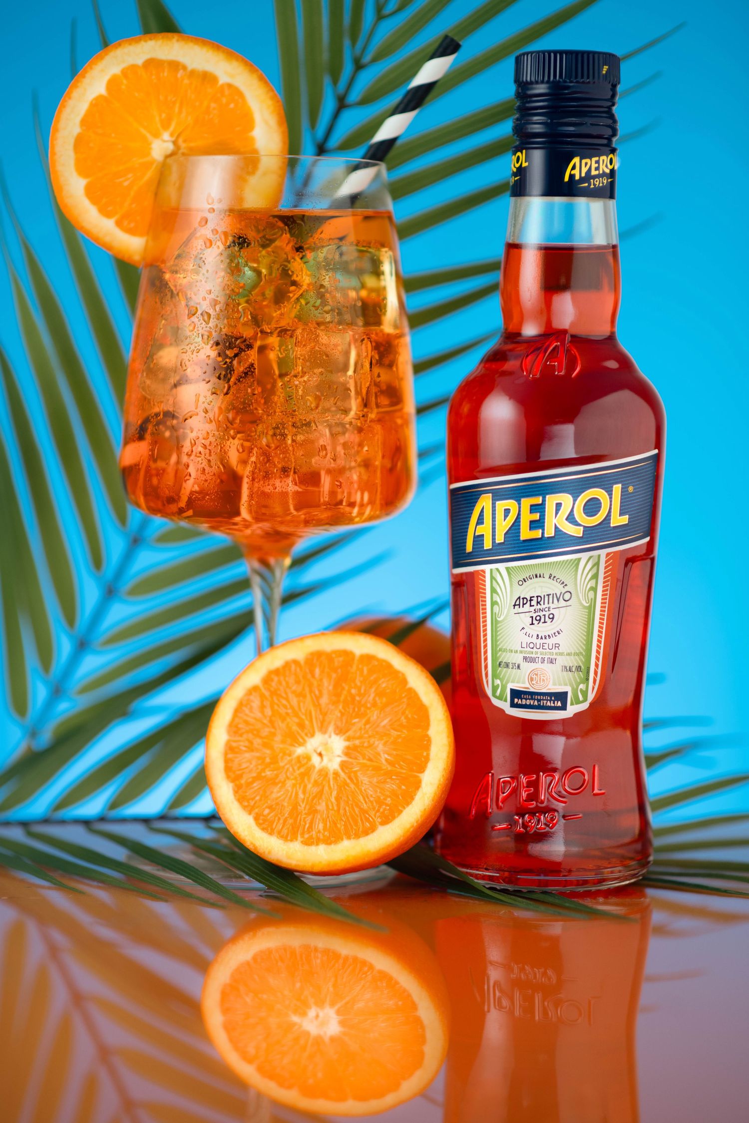 Aperol Spritz cocktail with orange slices next to an Aperol bottle on a blue tropical background.