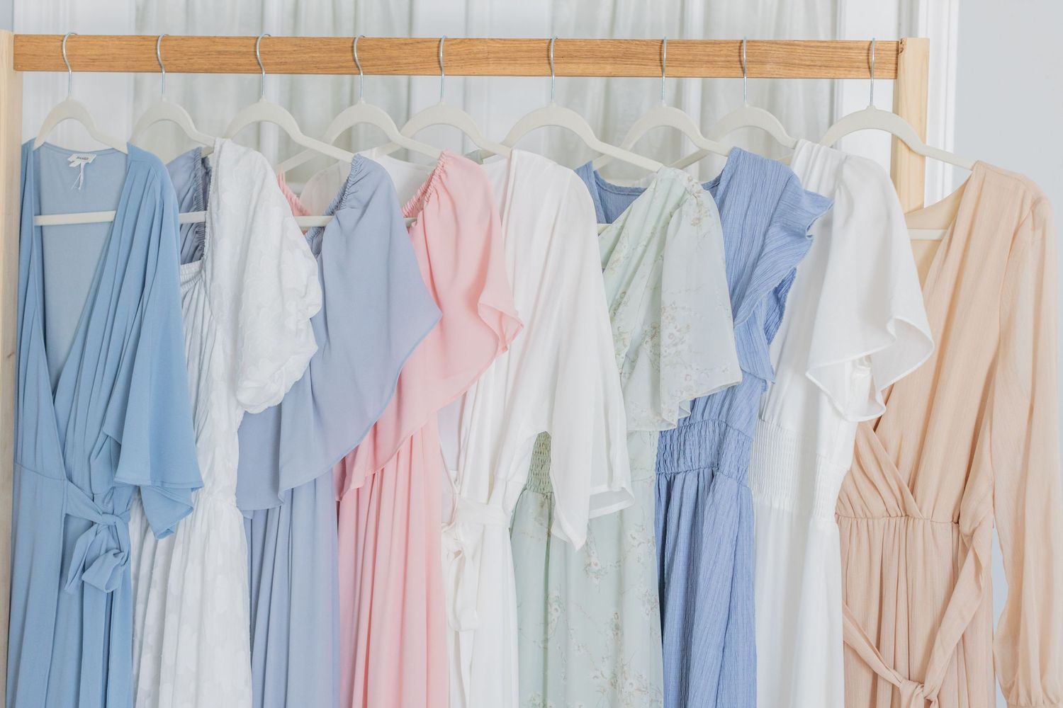 A row of pastel blue, pink, and cream colored bridesmaid robes hanging on a wooden hanger against a white wall.