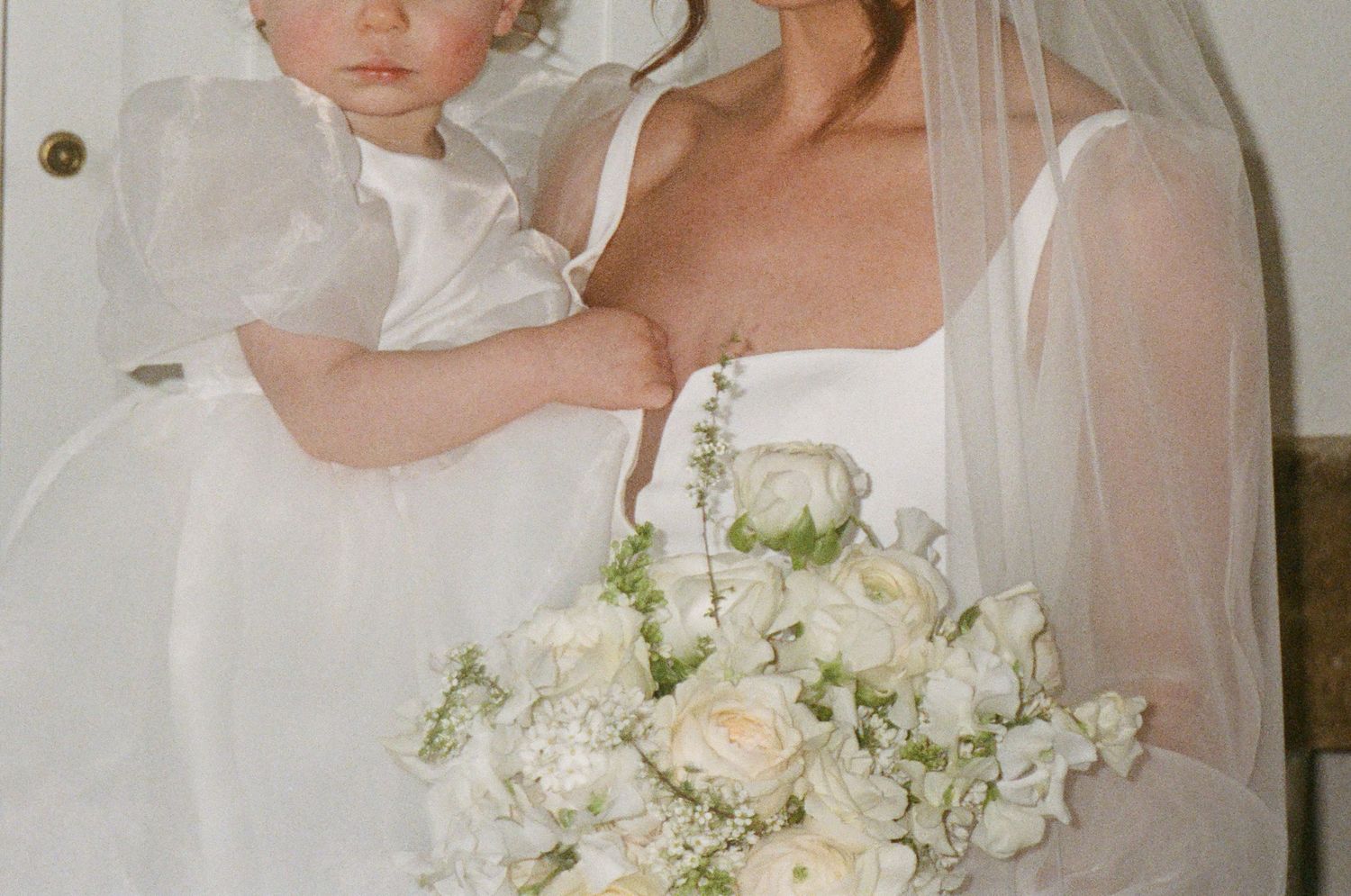 A beautiful white wedding bouquet held by a bride in a sheer veil alongside a little child.