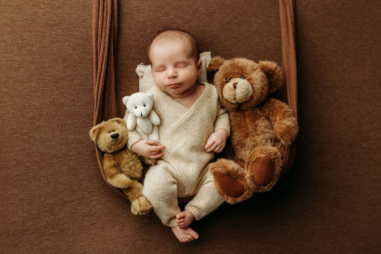 Newborn baby sleeping peacefully in a hammock surrounded by three teddy bears on a brown background.
