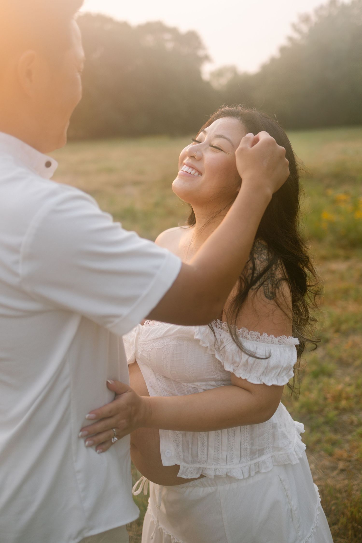 Couple in white outfits embracing in a sunlit field at golden hour with trees in the background.