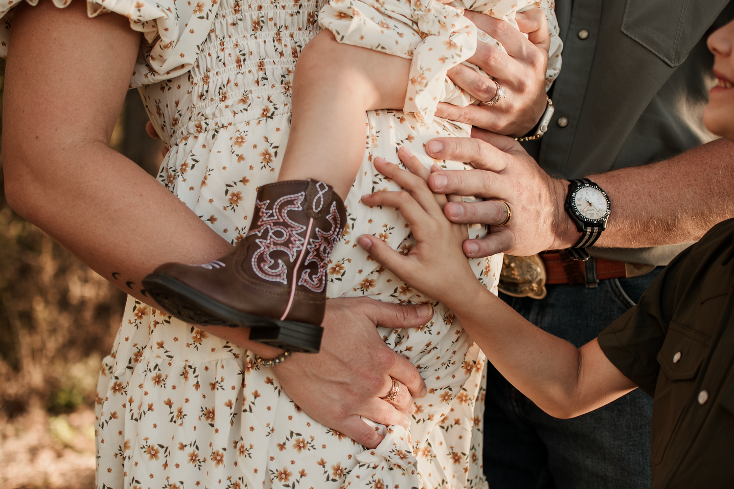 Couple holding tiny cowboy boots and clasped hands, pregnancy announcement with floral dress and leather watch.