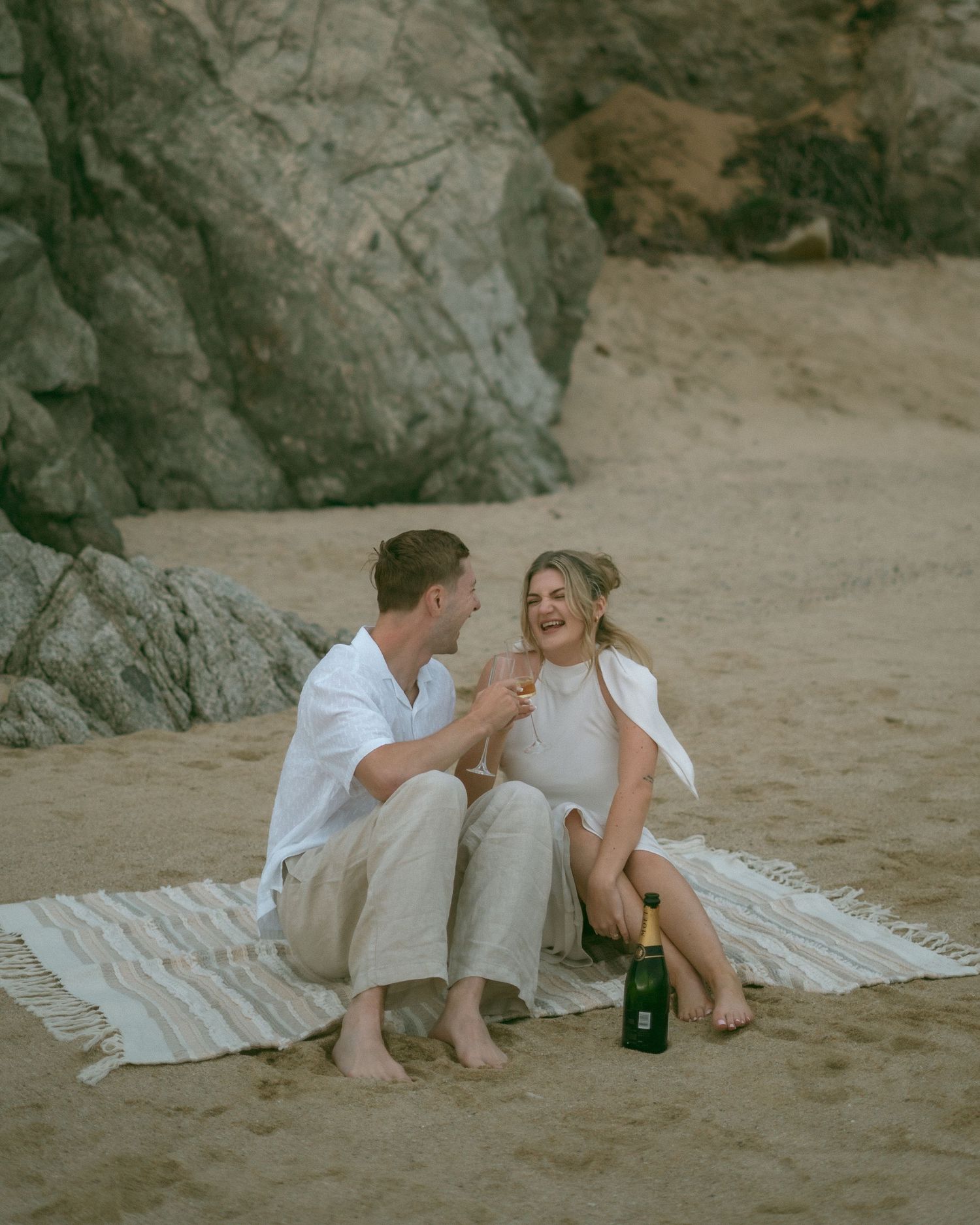 A couple in white attire toasting with wine glasses on a beach at sunset, woman with tattoos.