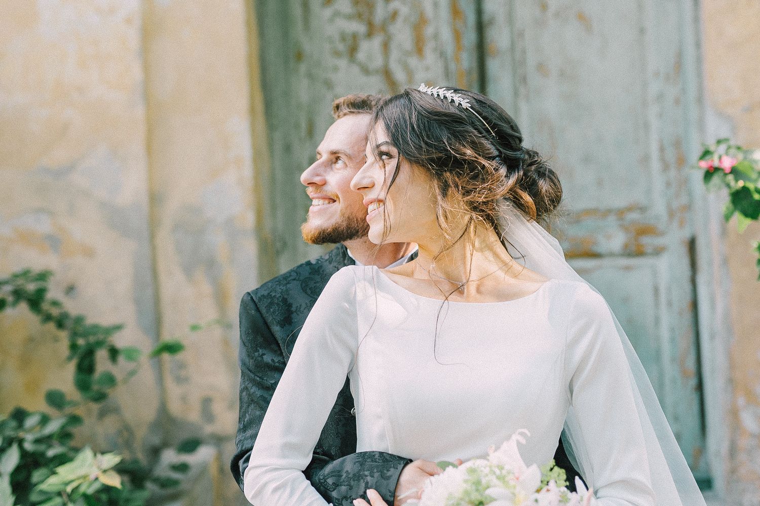 A bride and groom share a romantic moment outdoors, with the bride in a white gown holding a bouquet near a rustic door.